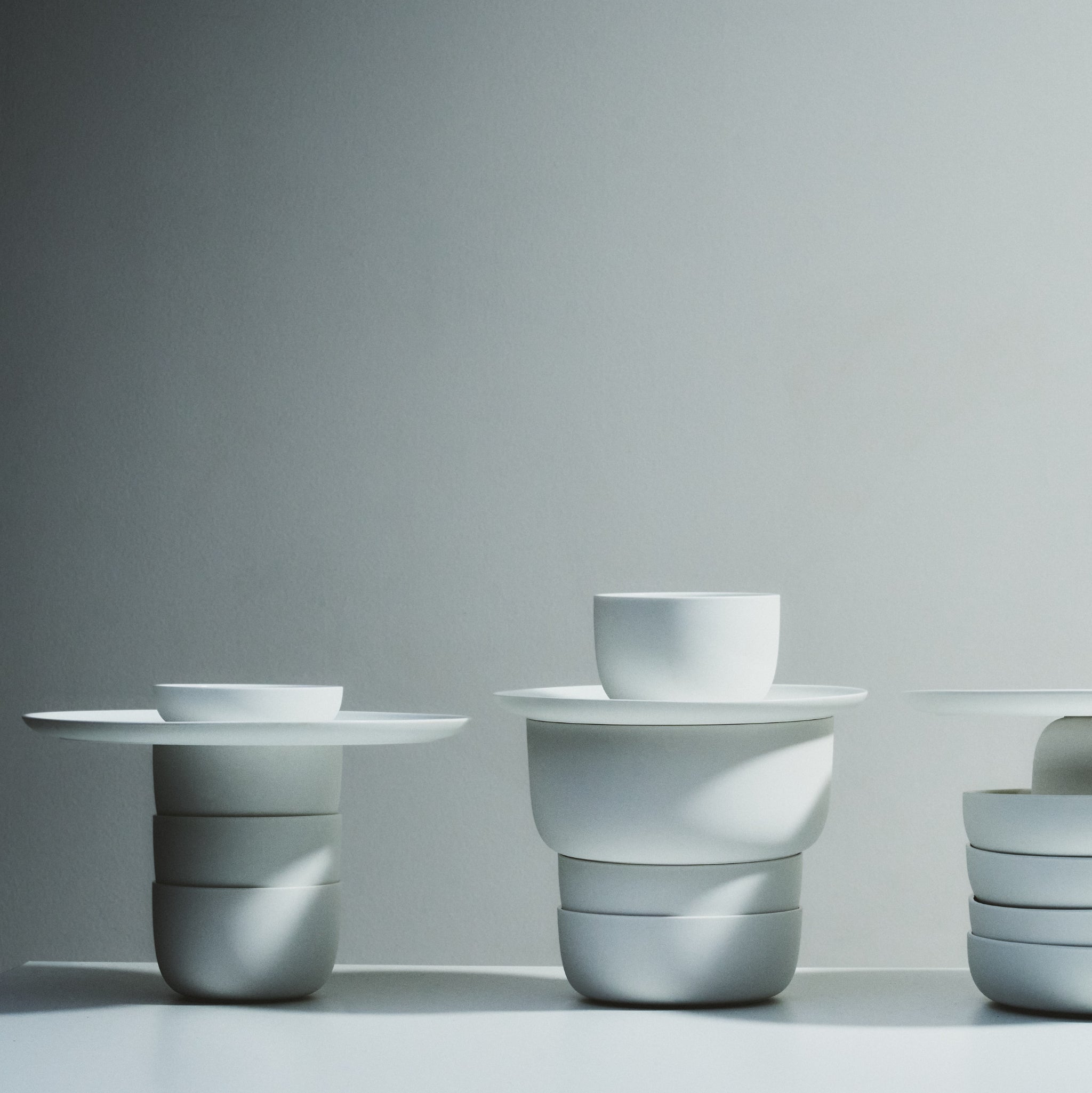 Minimalistische uitstalling van Serax Base Diepe Kom D20 keramische kommen, gestapeld in evenwichtige geometrische vormen op een strak oppervlak. Zachte verlichting werpt zachte schaduwen tegen een effen, lichtgrijze achtergrond, die de moderne Serax stijl vastlegt.