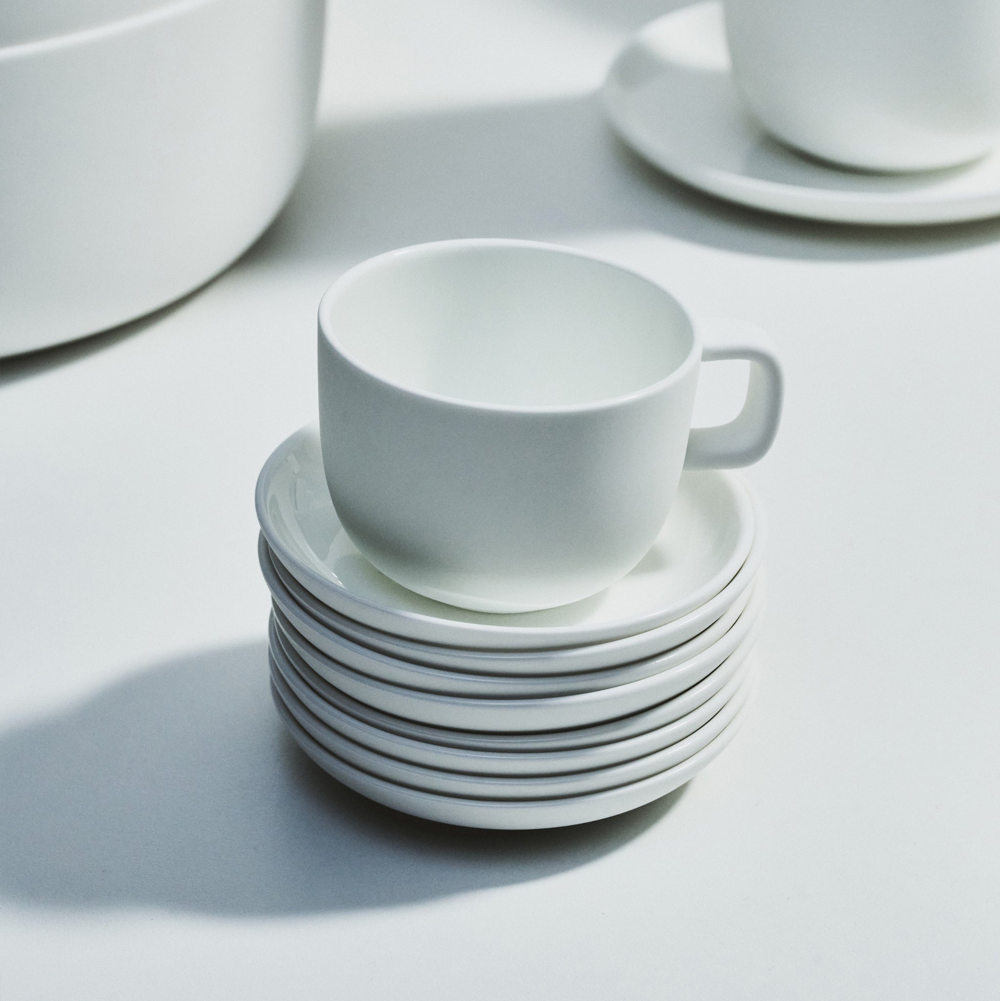 Een Serax Base Espresso Kop met Oor 10 cl rust op een stapel van zes bijpassende schotels, terwijl een andere kop en schotel set gedeeltelijk zichtbaar is daarachter. Zachte schaduwen en helder licht accentueren het elegante Serax ontwerp op een wit oppervlak.