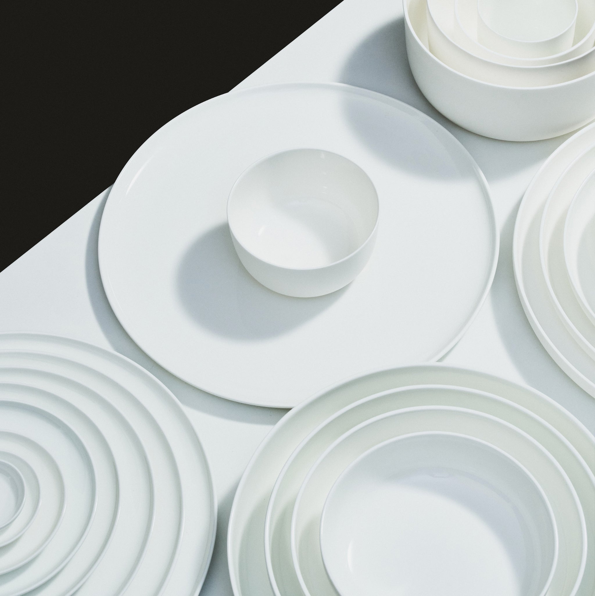 Een keurig gerangschikte set van Serax Base Hoge Kom D12 witte keramische kommen en borden rust op een wit oppervlak, van bovenaf gezien met een vleugje zwart in een hoek, wat de verfijnde stijl van Serax oproept.