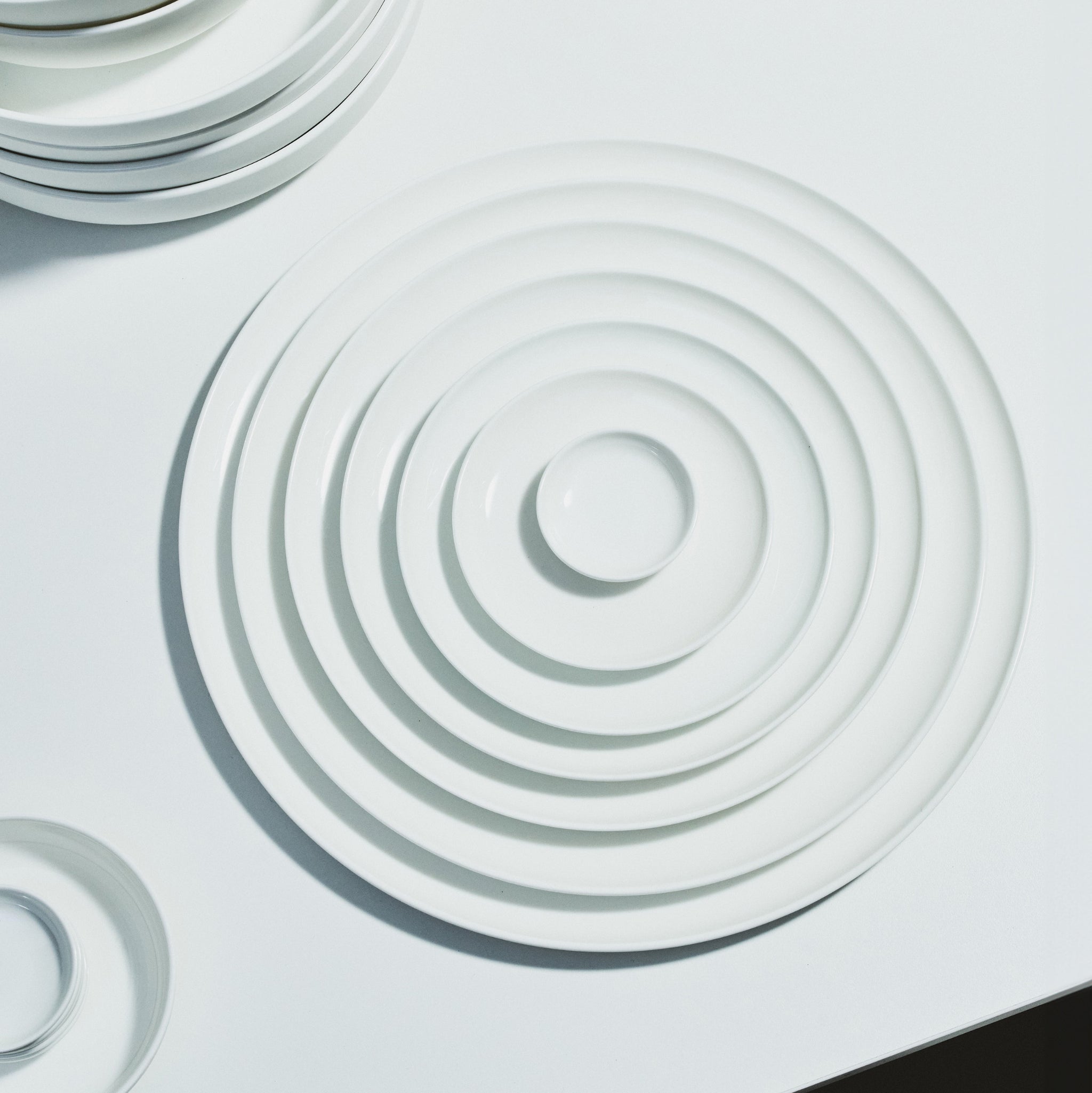 Een stapel Serax Base Laag Bord D16 borden van Serax, gemaakt van wit porselein, is concentrisch gerangschikt op een wit oppervlak, waardoor een rimpeleffect ontstaat. De verlichting benadrukt hun elegante ronde vormen.