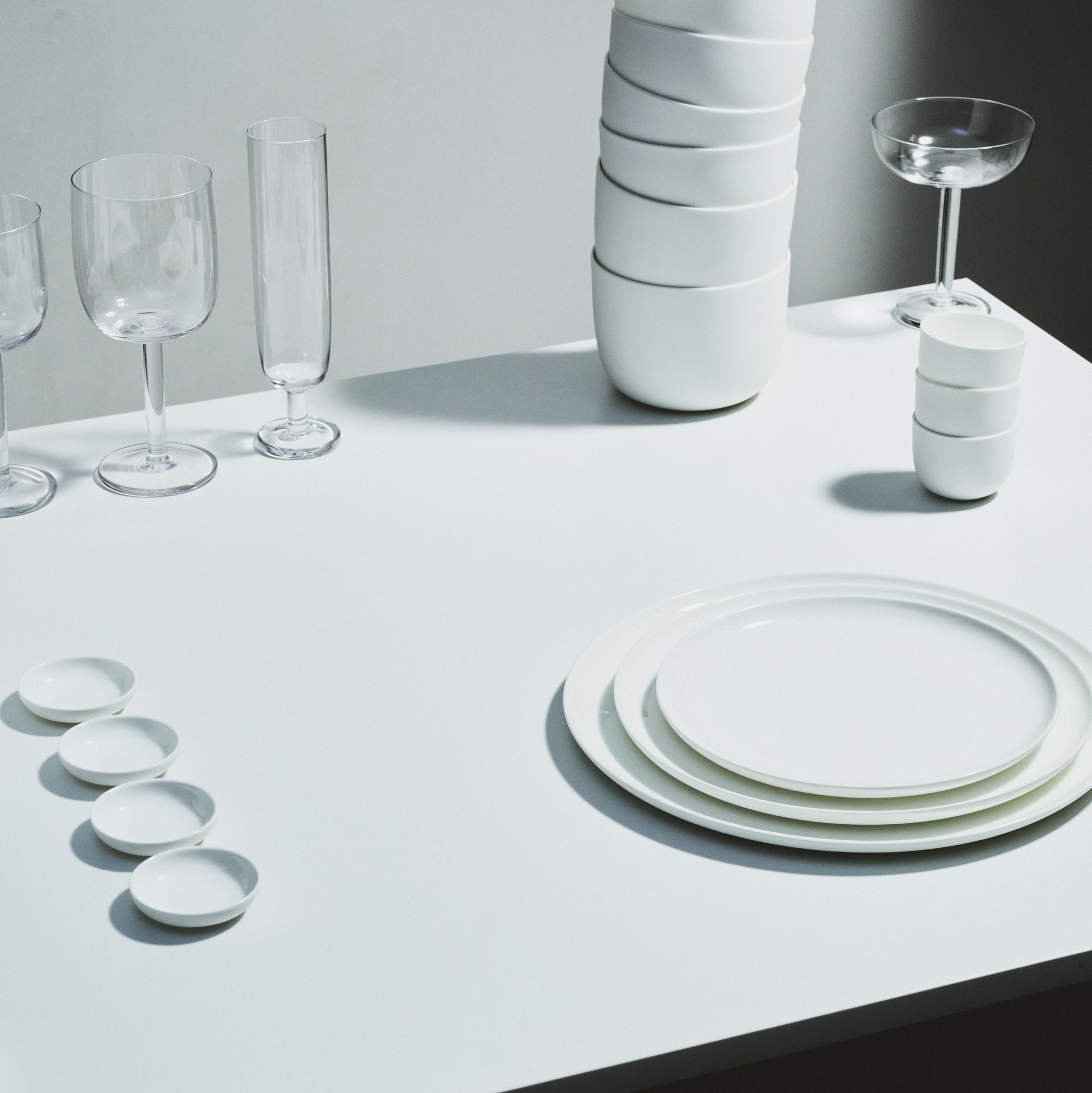 Een minimalistische tafelschikking met gestapelde Serax Base Laag Bord D8 borden en kleine schaaltjes van Serax, plus vier lege glazen - netjes gerangschikt op een wit oppervlak met zachte verlichting die zachte schaduwen werpt.