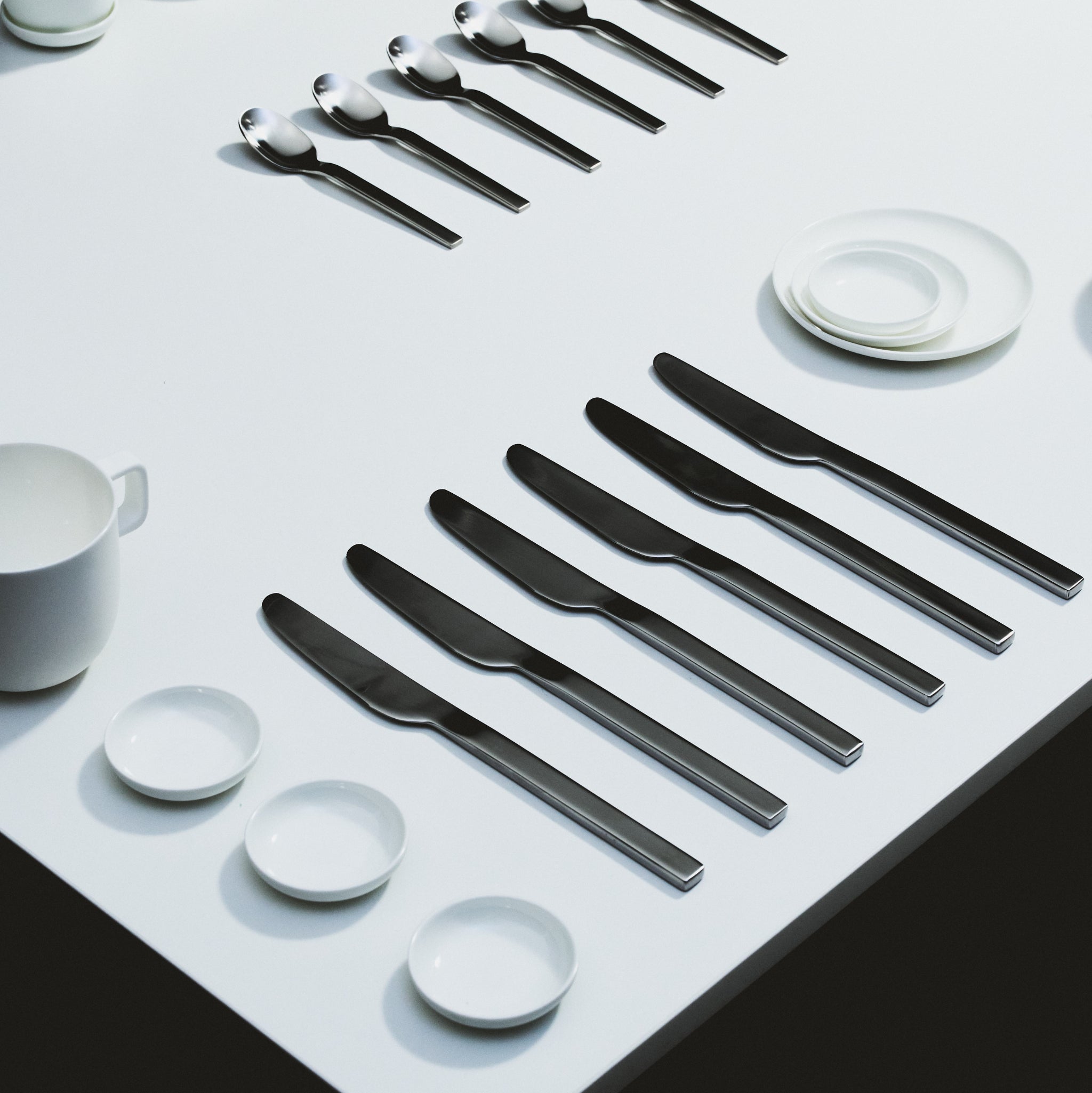 Een minimalistische tafelschikking op een wit oppervlak met Serax Base Laag Bord D8 borden van Serax, met zes roestvrijstalen messen, lepels en kleine witte kommen, schotels en kopjes die netjes in parallelle rijen zijn geplaatst.