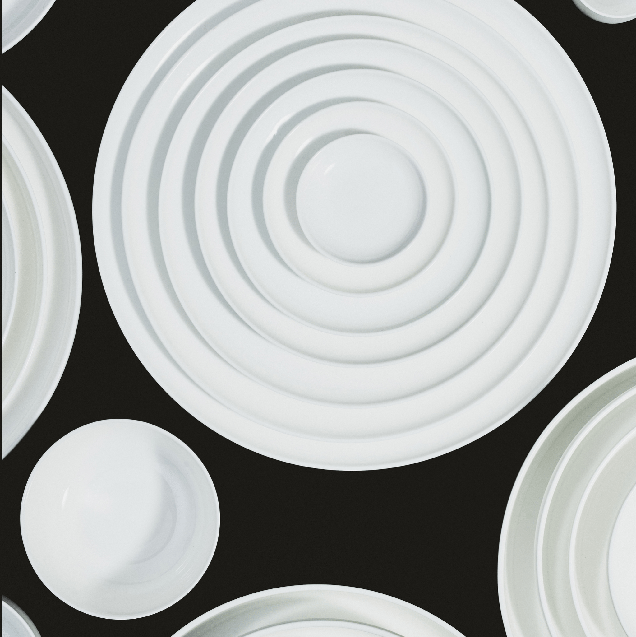 Serax Base Theekop zonder Oor 35 cl kopjes van Serax zijn gerangschikt op een zwart oppervlak - sommige gestapeld in concentrische cirkels - en vormen een geometrisch, minimalistisch patroon met wit keramisch servies.