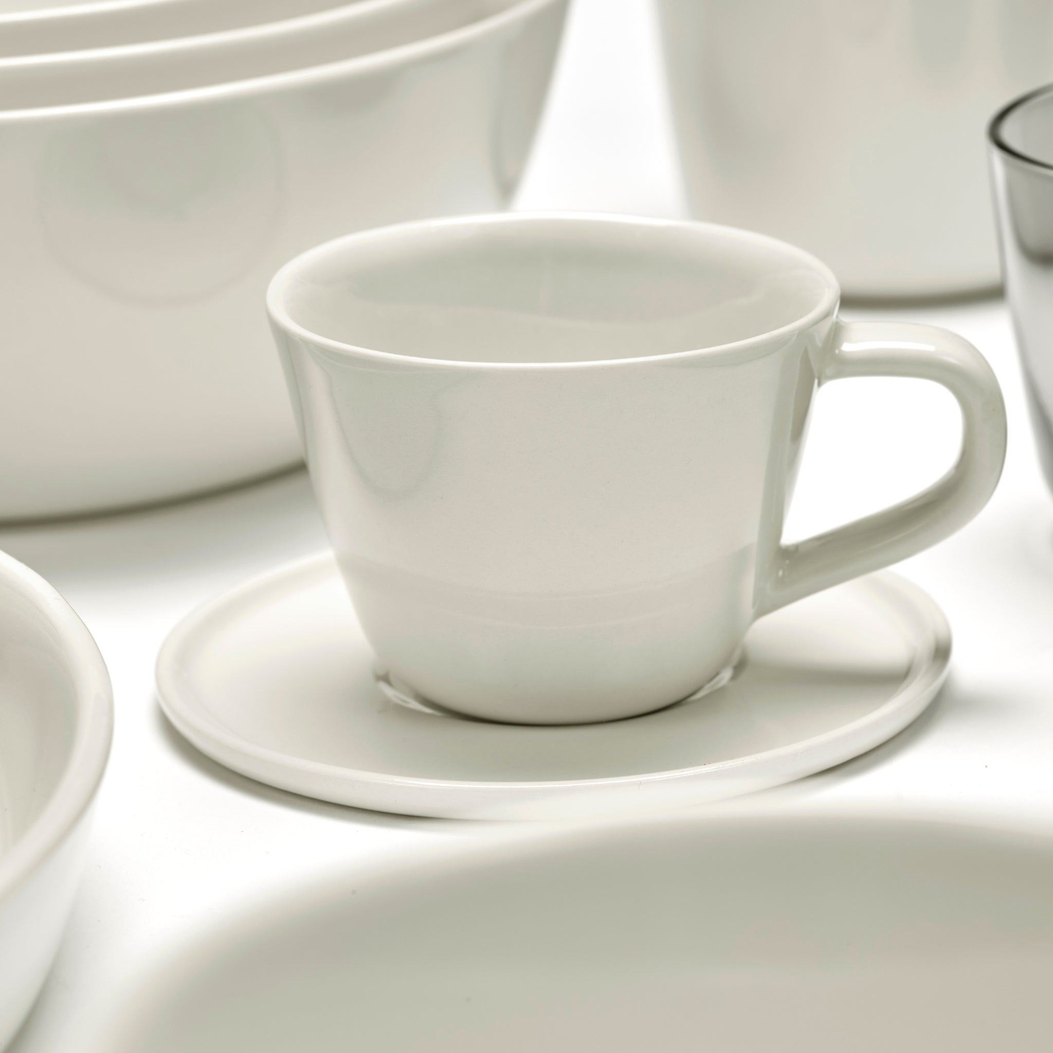 De Serax Cena Espresso Kop met Oor 12 cl staat op een bijpassende schotel, omringd door andere witte schalen en kommen, allemaal gerangschikt op een wit oppervlak.