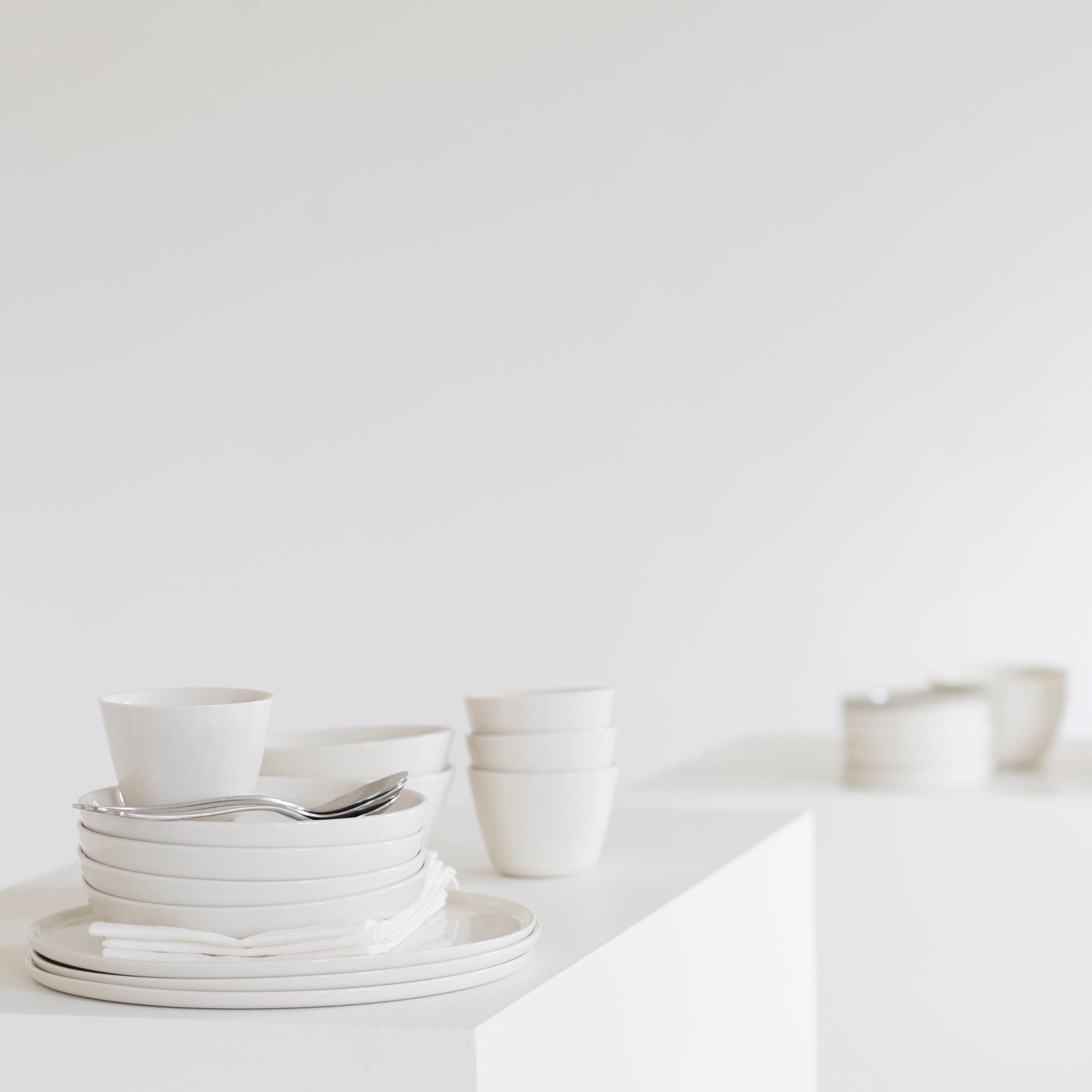 Een Serax Cena Theekop met Oor 37 cl van Serax staat netjes tussen ander wit servies op een wit oppervlak, tegen een minimale lichte achtergrond met meer wit servies in de verte.