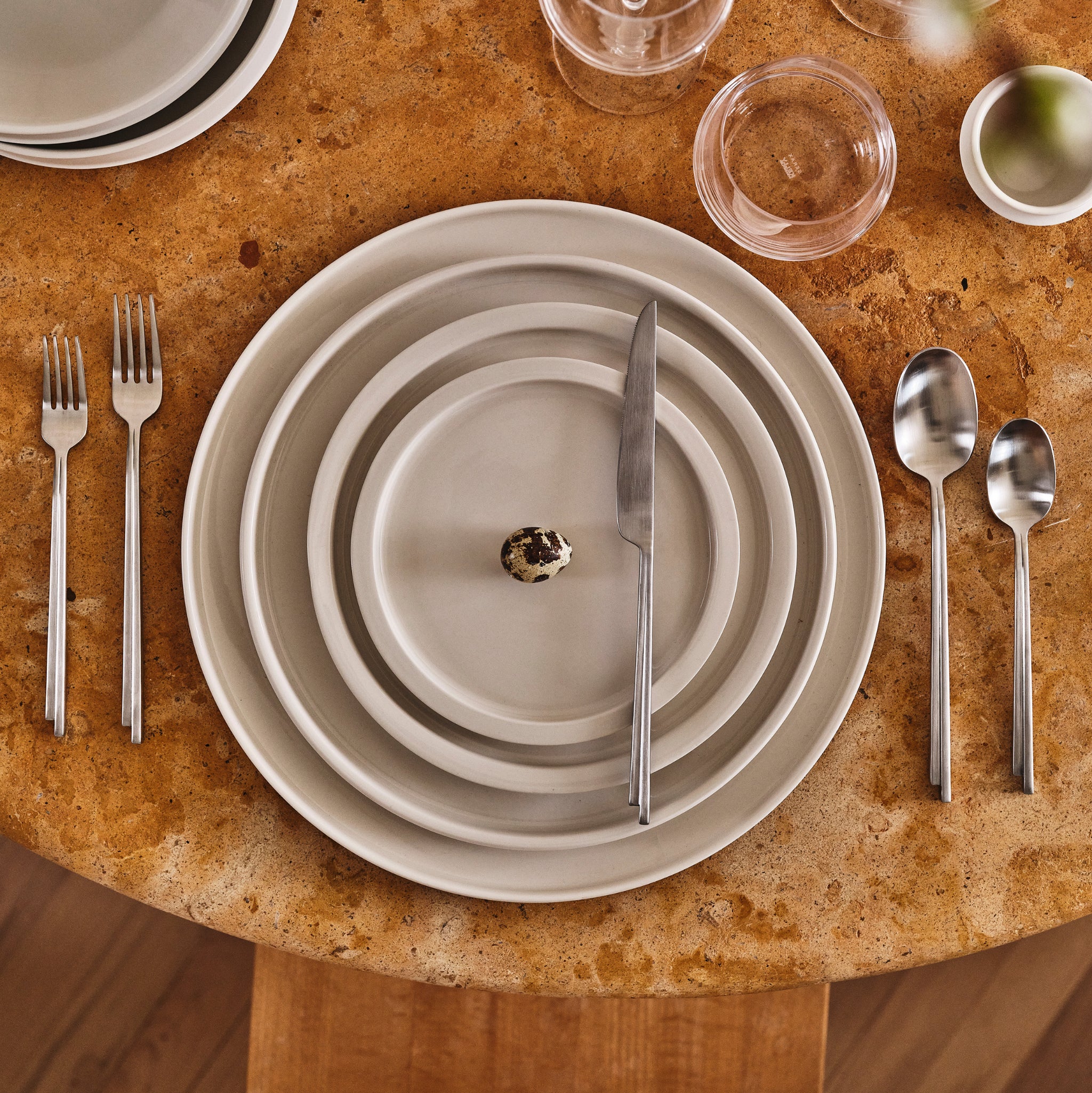 Een formele tafelschikking op een ronde, bruine stenen tafel is voorzien van gestapelde Serax Dune Laag Bord Alabaster D33 borden, een klein kwarteleitje in het midden, netjes gerangschikt bestek en elegant glaswerk.