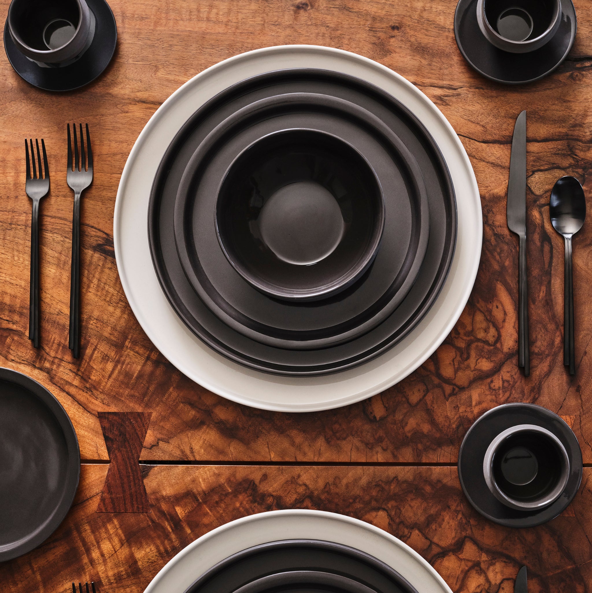 Een houten tafel is gedekt met gestapelde Serax Dune Laag Bord Slate D17 borden, kommen en kopjes op witte porseleinen borden. Zwart bestek maakt de moderne, symmetrische opstelling, geïnspireerd door Kelly Wearstler, compleet.