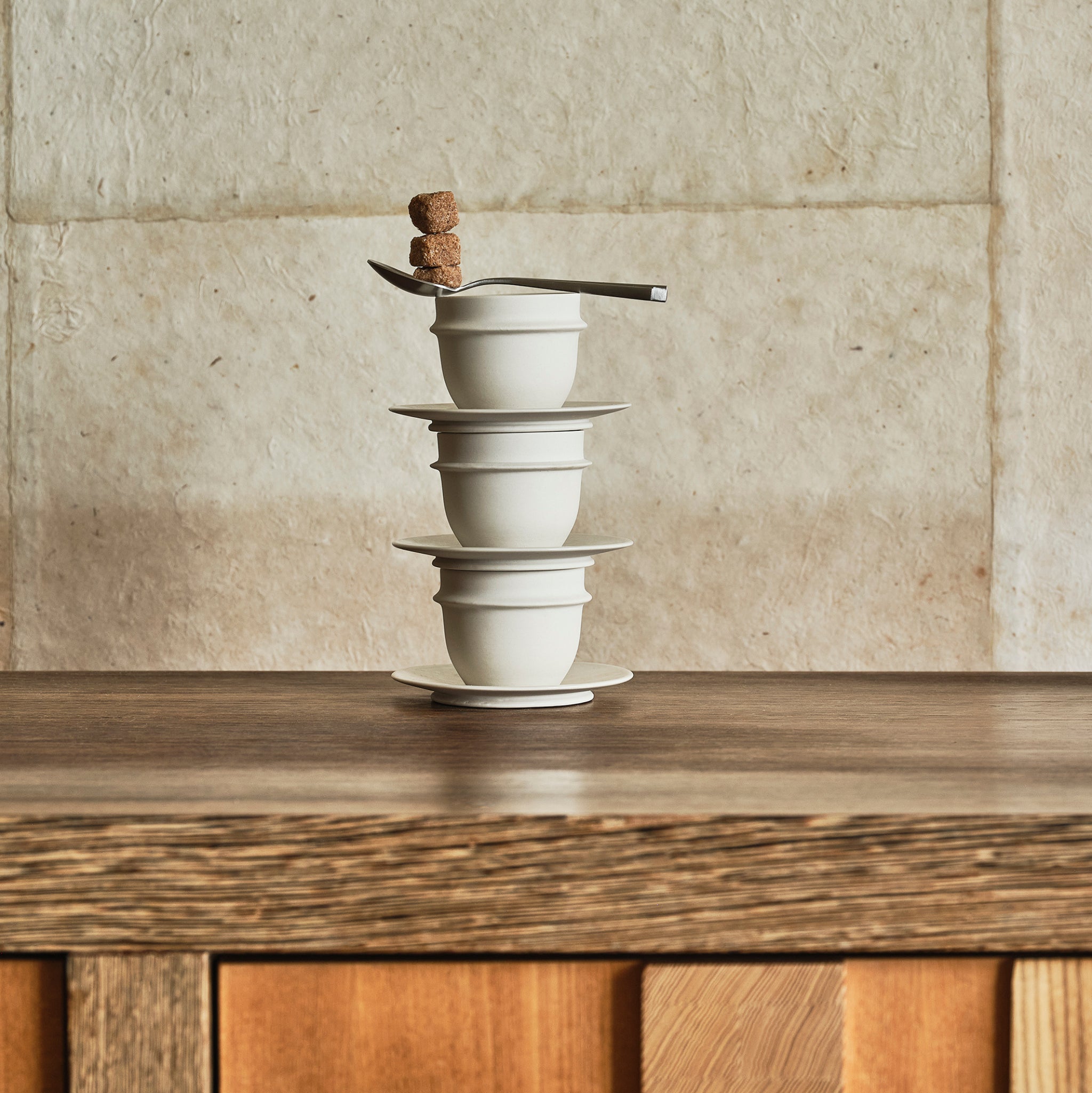Een stapel van drie witte Serax Dune Schotel Espresso Kop Albast D11 kopjes met schotels staat op hout, met daarop een lepel en bruine suikerklontjes. Het tafereel speelt zich af tegen een lichte stenen muur.
