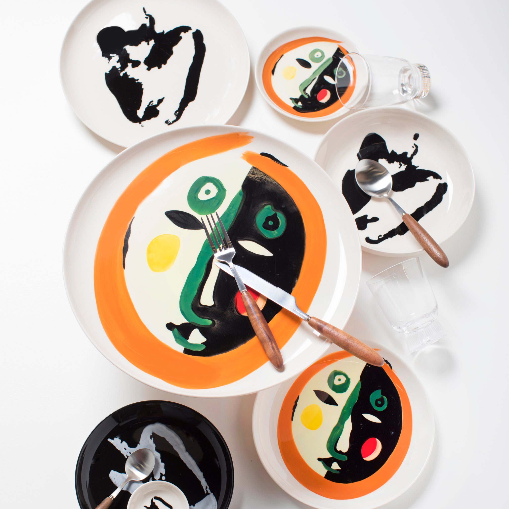 Een levendige tafelsetting met de keramische borden „Serax Feast Ottolenghi Laag Bord Face N°1 D16“ van het merk Serax, die zich onderscheiden door opvallende abstracte ontwerpen in oranje, groen, geel en zwart, geïnspireerd op gezichten, gecombineerd met glazen en bestek op een strak wit oppervlak.