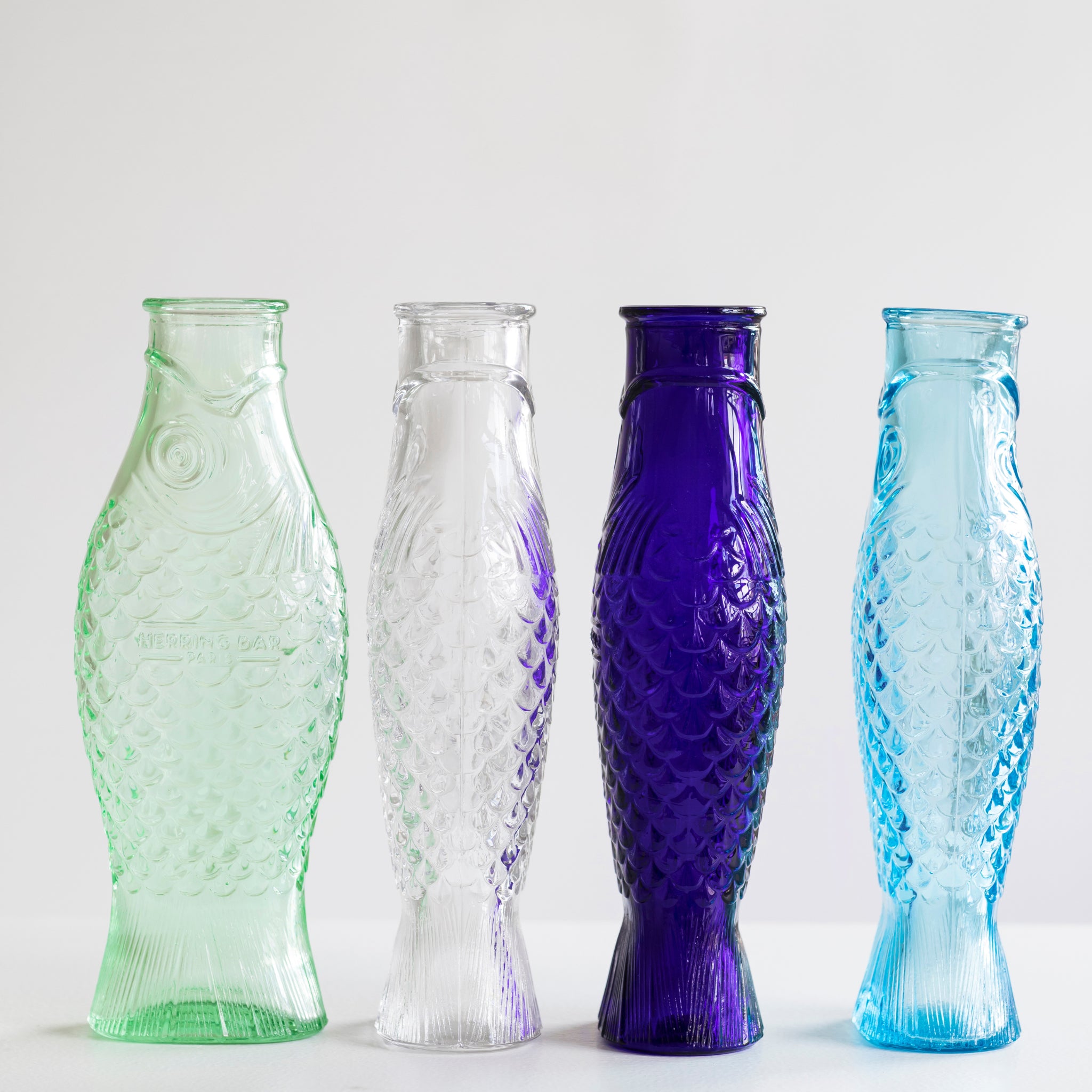 Vier glazen flessen van Serax Fish & Fish Karaf Helder 85 cl, ontworpen door Paola Navone voor Serax, staan op een rij. Elke fles heeft getextureerde schubben en is verkrijgbaar in lichtgroen, helder, donkerblauw en lichtblauw tegen een witte achtergrond.