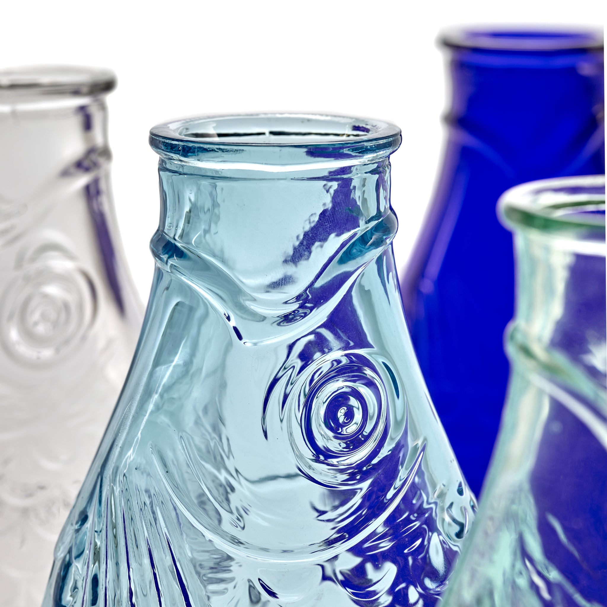 Close-up van vier Serax Fish & Fish Karaf flessen (85 cl) in lichtblauw, kobalt en helder glas, elk met gedetailleerde schubben en ogen. Ontworpen door Paola Navone voor Serax, weergegeven tegen een witte achtergrond.