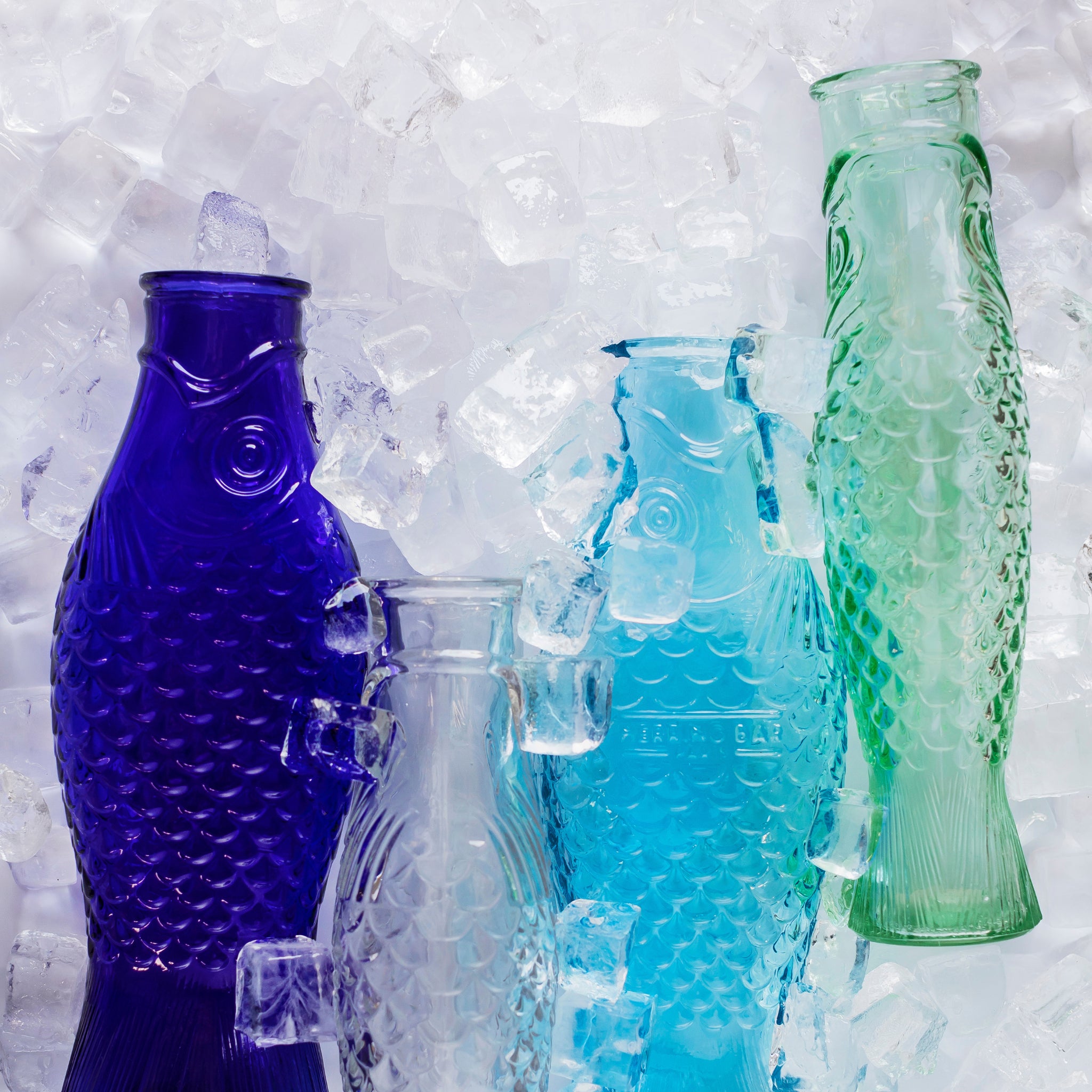 Vier Serax Fish & Fish Karaf Lichtblauw 85 cl flessen, gemaakt door Serax, staan rechtop op ijs in blauwe, groenblauwe en groene tinten. Het getextureerde glas en de grillige visvorm komen prachtig uit tegen een kraakheldere witte achtergrond.