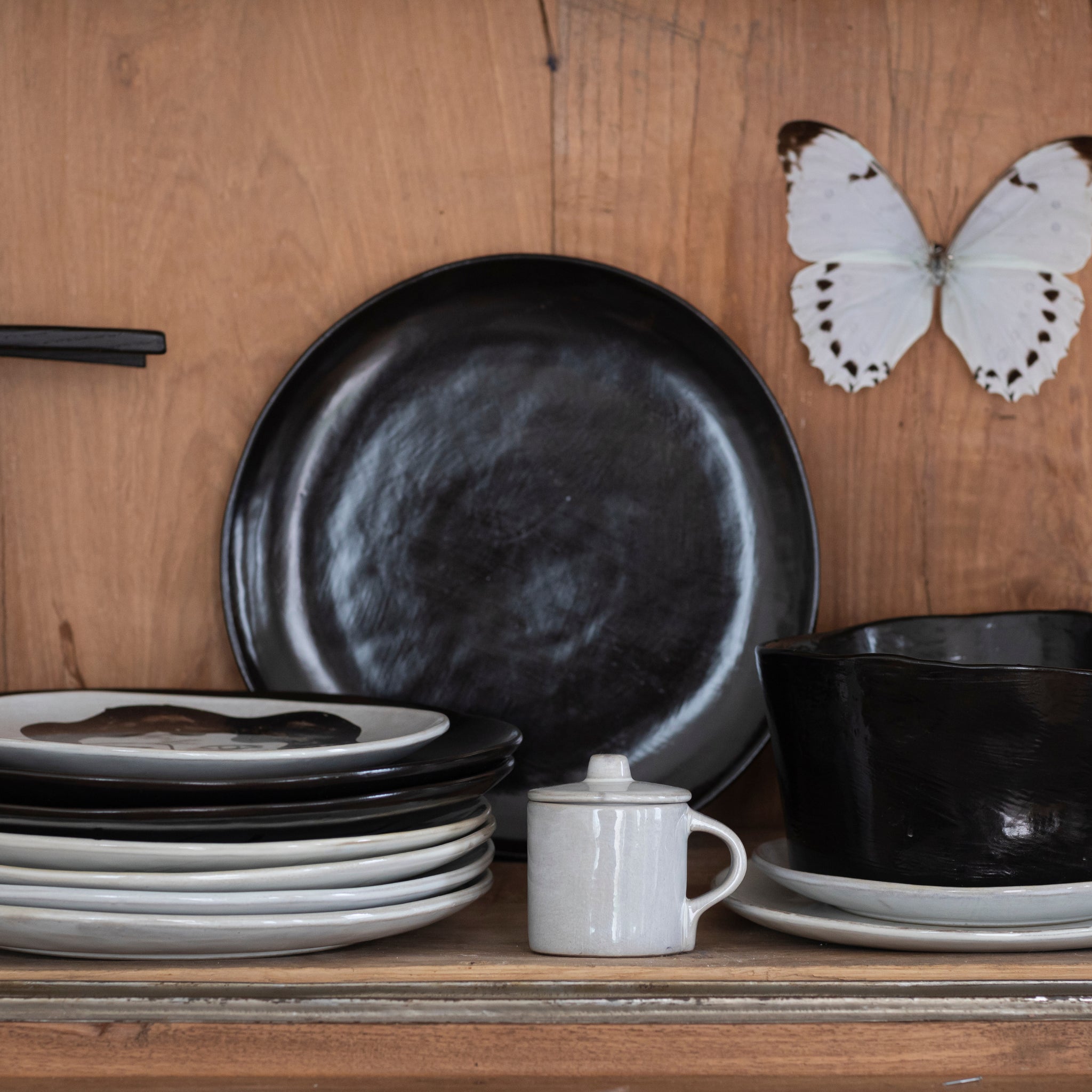 Op een houten plank staat de Serax La Mère Serveerschaal Ebony D31, gestapeld zwart en wit keramiek, een kleine witte pot met deksel, een zwarte kom en een opgezette vlinder - een elegant eerbetoon aan Serax La Mère van Marie Michielssen.