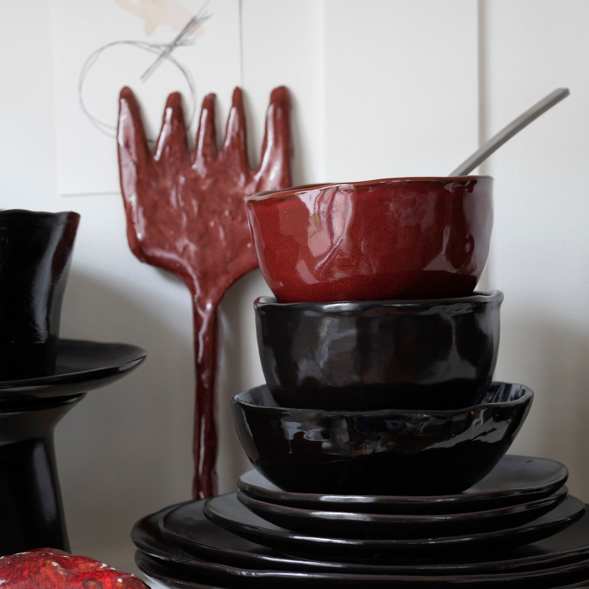 Een stapel glanzende keramiek kommen staat op borden naast de Serax La Mère Serveerschaal Ebony D37 van Serax, met een groot rood dienblad en lepel met textuur, allemaal geplaatst tegen een witte muur met minimale kunst.