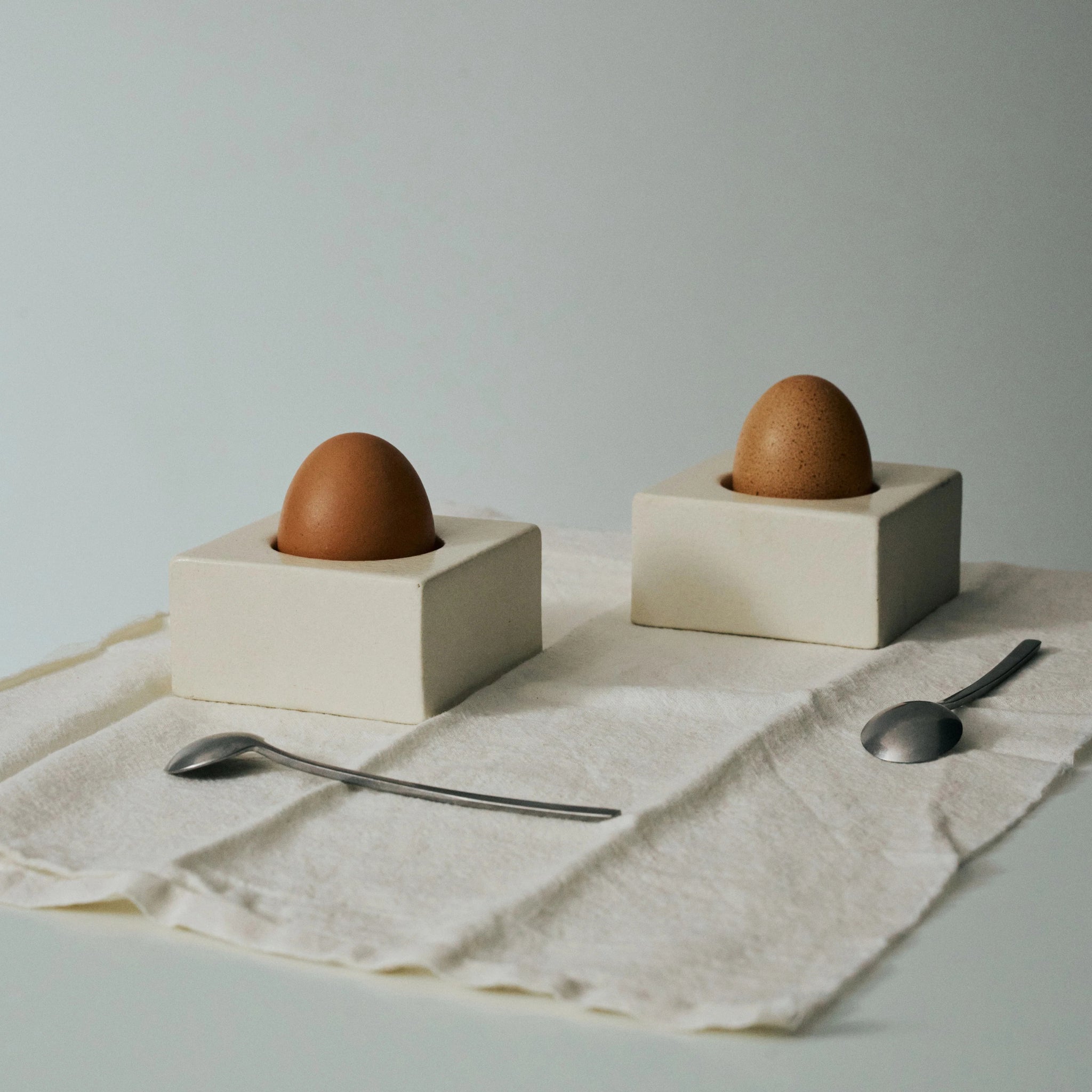 Twee bruine eieren zitten rechtop in Serax Uovo Eierdopje Wit van Serax, gemaakt van wit aardewerk, op een crèmekleurig kleed met twee metalen lepels ernaast. Het zacht verlichte, minimale tafereel benadrukt het eigentijdse ontwerp.