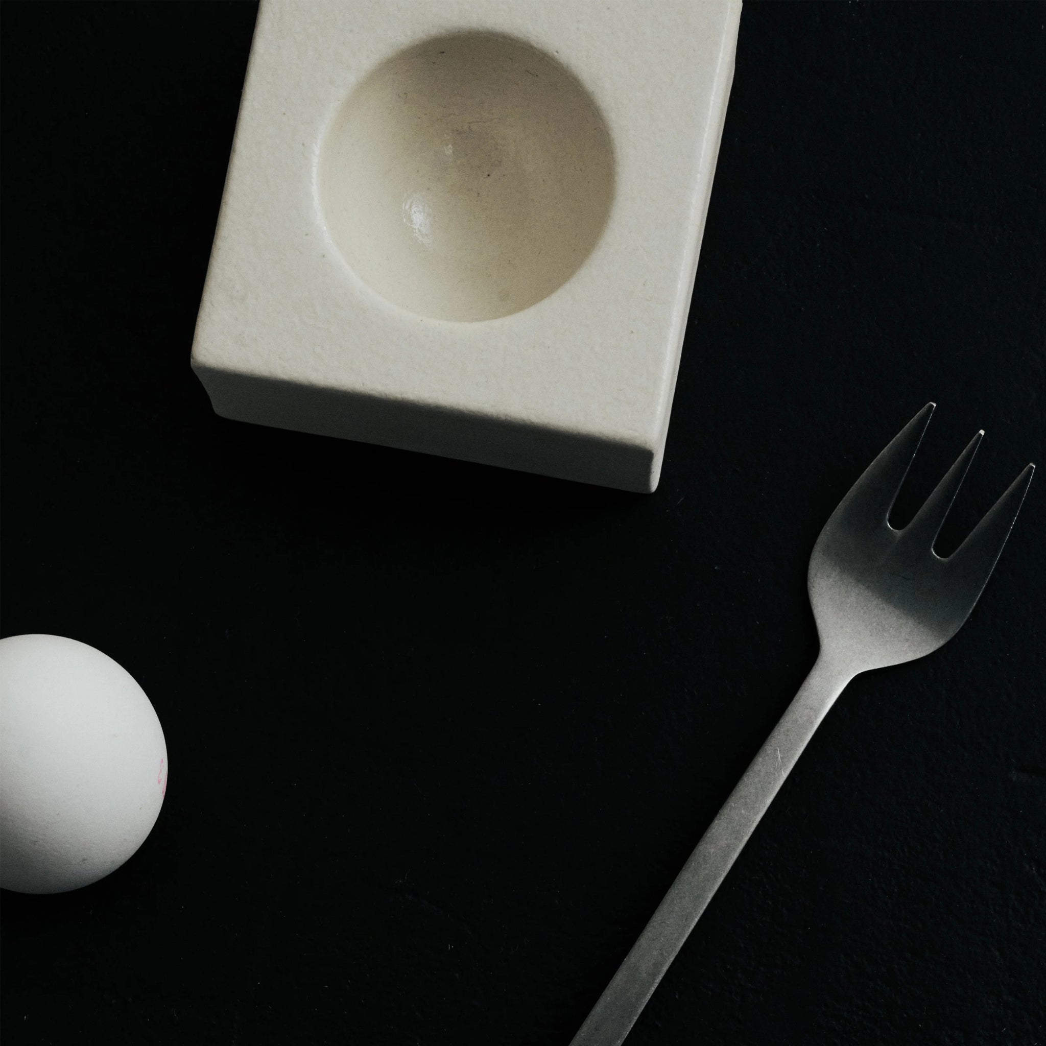 Een wit ei zit in een Serax Uovo Eierdopje Wit van Serax, gecombineerd met een metalen drietandige vork op een donkere, gestructureerde achtergrond voor een eigentijdse look.