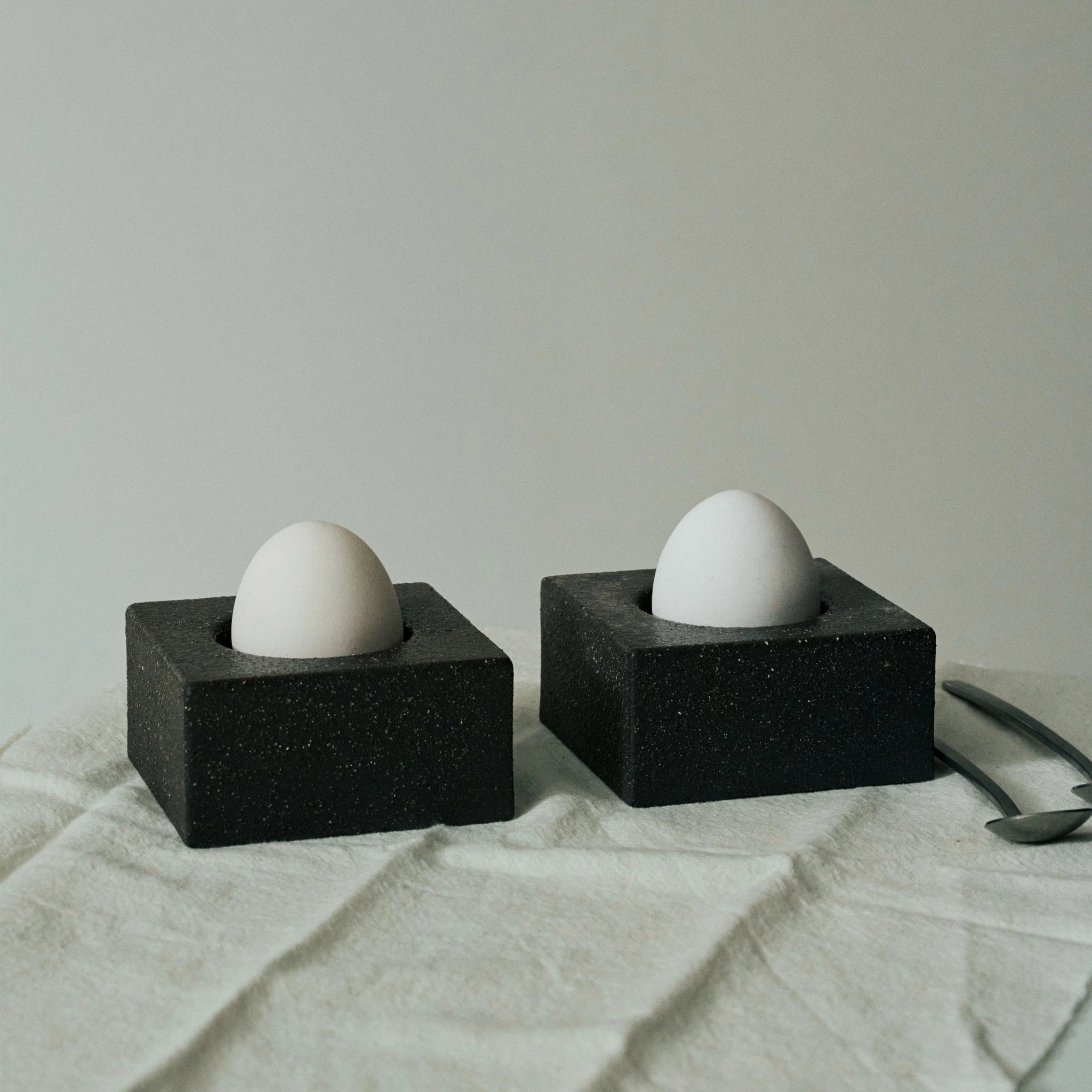 Twee witte eieren staan rechtop in de Serax Uovo Eierdopje Zwart eierdopjes van Serax op een licht, gestructureerd doek, met twee metalen lepels in de buurt. De achtergrond is effen en zacht verlicht.