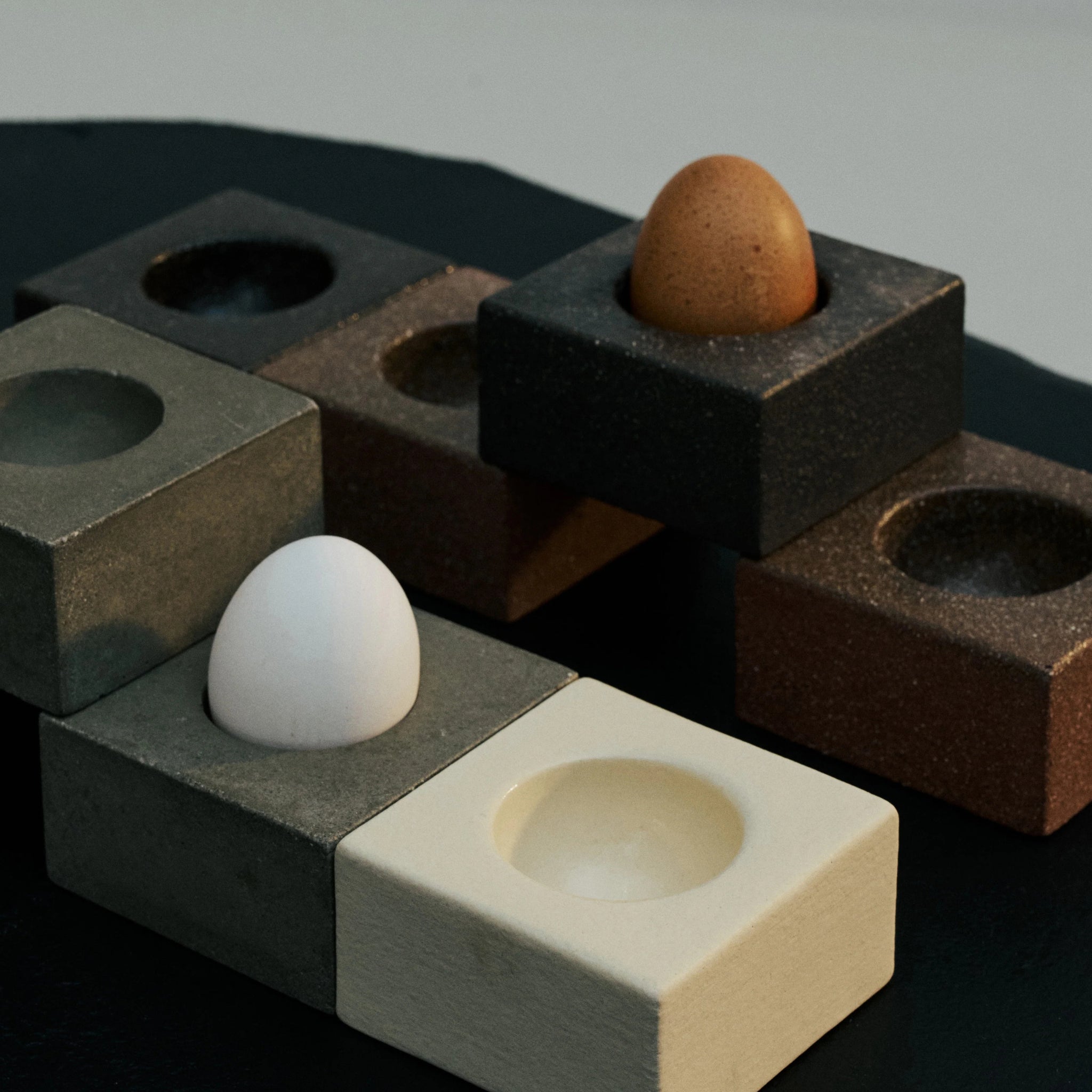 Een bruin en een wit ei zitten in rechthoekige Serax Uovo Eierdopje Zwart houders van Serax. Hun ronde inkepingen creëren een opvallend geometrisch patroon op het donkere oppervlak.