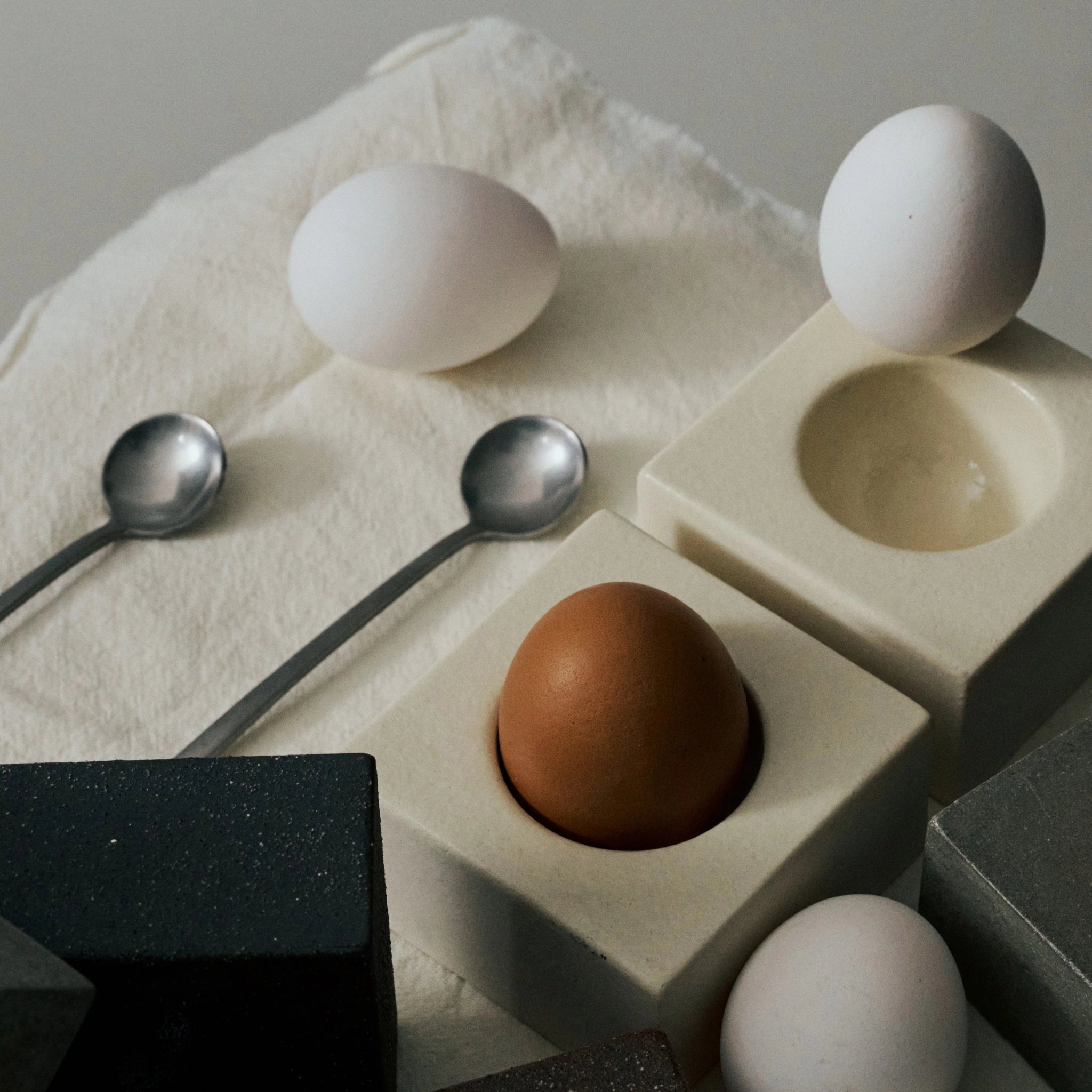 Drie eieren (twee witte, een bruine) liggen op een zacht wit doek met twee metalen lepels en het Serax Uovo Eierdopje Zwart, waardoor een moderne compositie ontstaat in de stijl van Catharina Bossaert ontwerp.