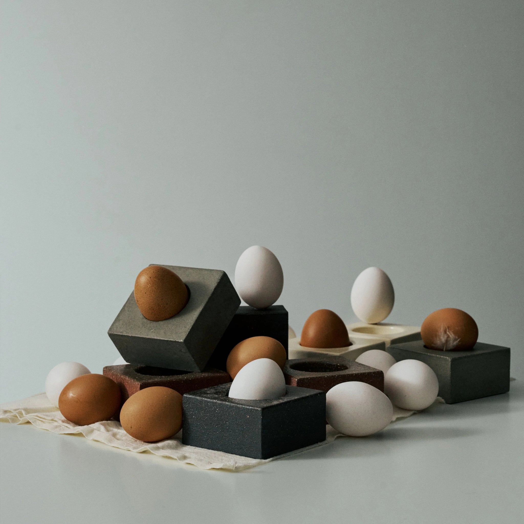 Bruine en witte eieren, sommige in het Serax Uovo Eierdopje Zwart van Serax, zijn gerangschikt op grijze en beige geometrische blokken op lichte stof tegen een effen, lichte achtergrond.