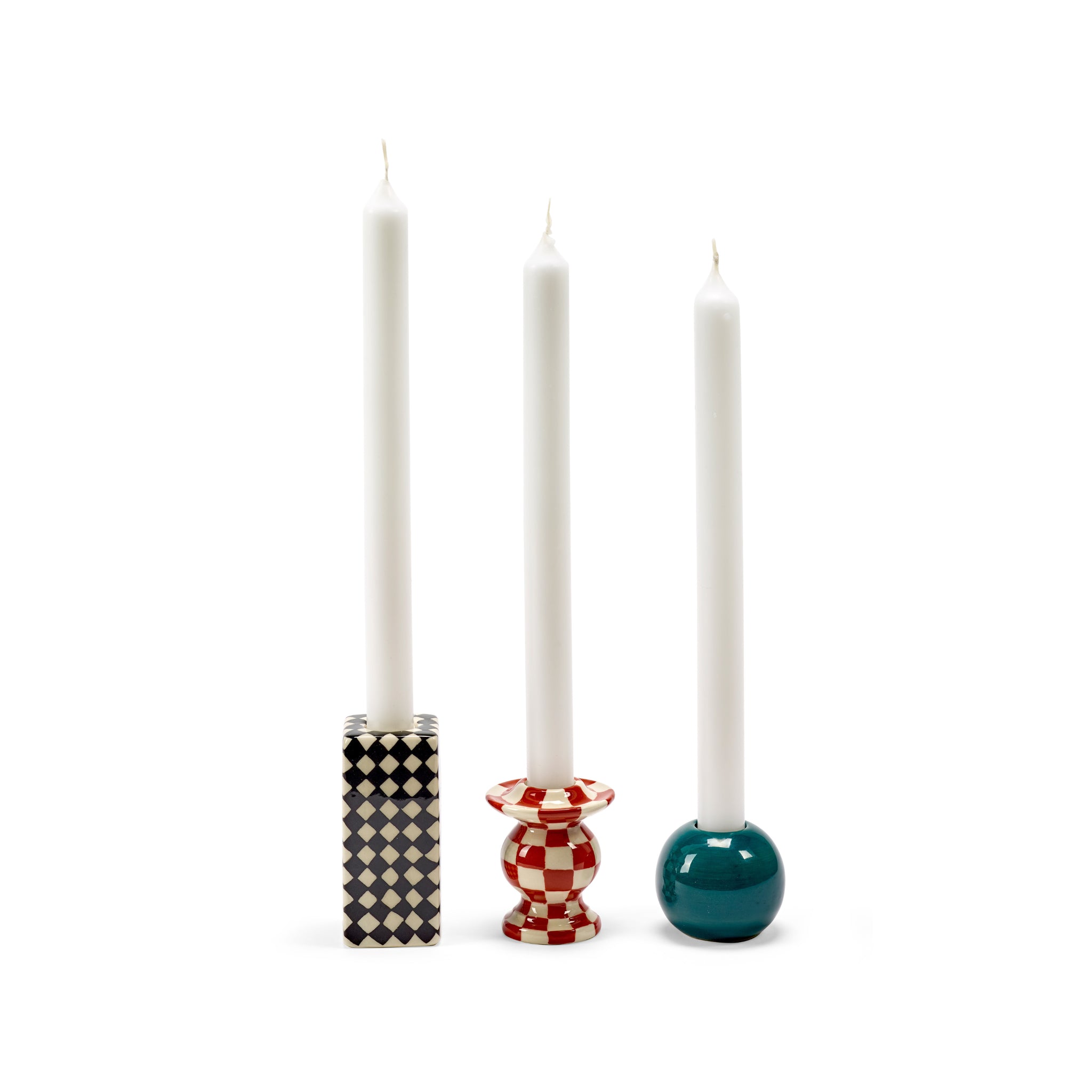 Drie witte kaarsen in Serax Wouters & Hendrix Schaakbord Kandelaars houders: één met een zwart-wit geblokte vierkante voet, één met een rood-wit geblokte ronde voet en één met een glanzend groenblauwe ronde voet.