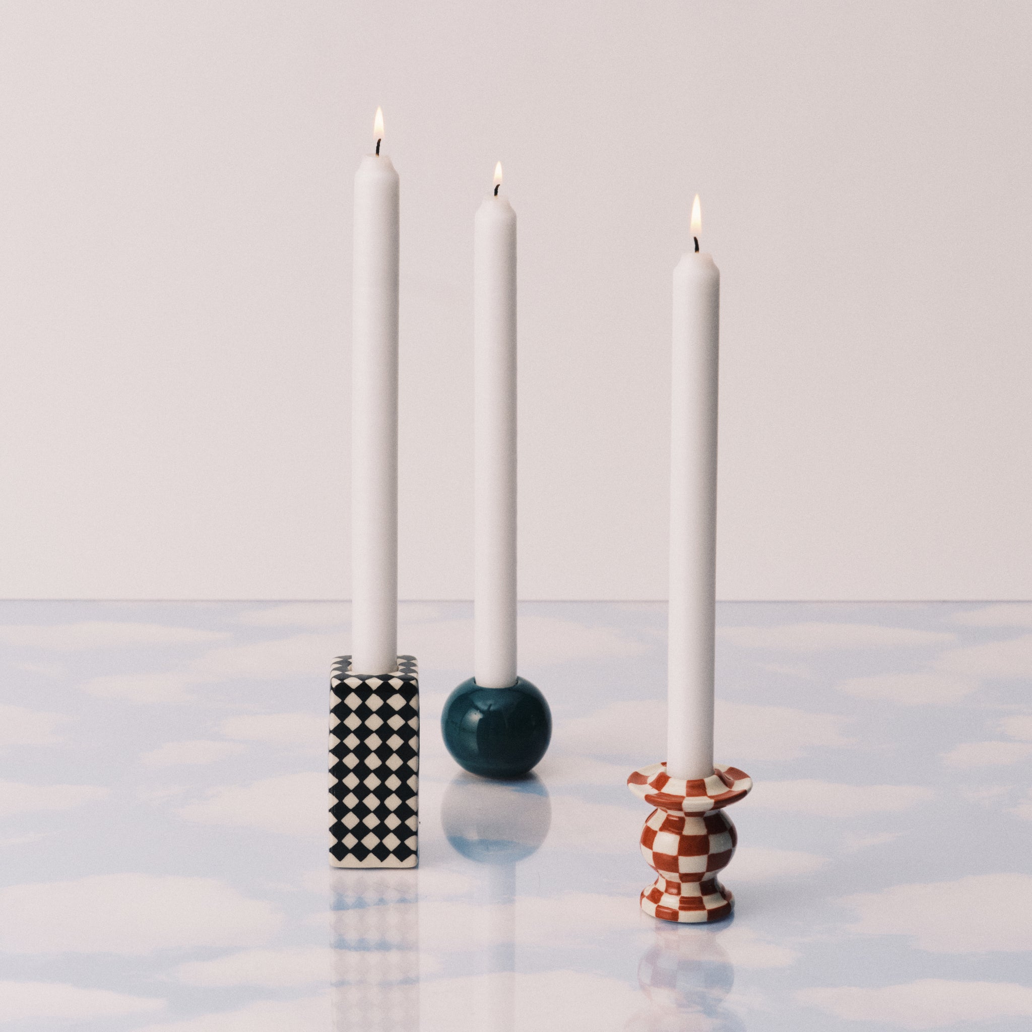 Drie brandende witte kaarsen in geometrische houders, waaronder een Serax Wouters & Hendrix Chessboard Kandelaars 3 st. van Serax, een groenblauwe bol en een oranje-witte voet, staan op een oppervlak met wolkenpatroon.