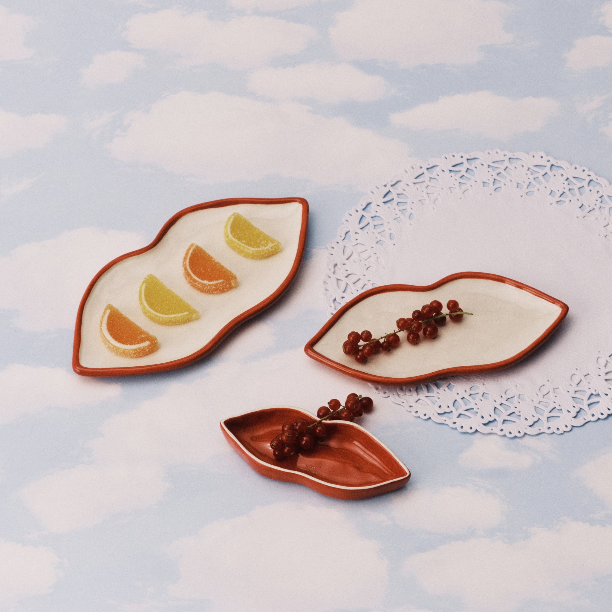 Drie Serax Wouters & Hendrix Lips Schalen 3 st. keramische borden van Serax staan op een oppervlak met wolkenpatroon; op een ervan liggen partjes citrusvruchten met een suikerlaagje, op de andere rode bessen, met een wit papieren kleedje op de achtergrond.
