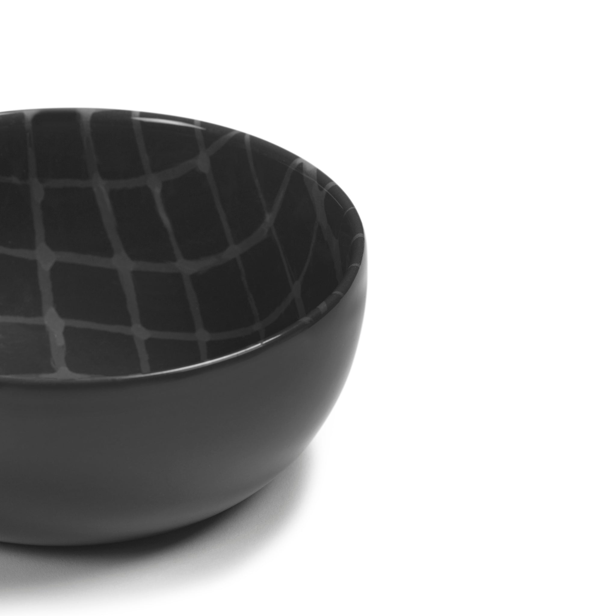 De Serax Zuma Kom Pacific D12,5, een kom van zwart keramiek van Serax met een op Bauhaus geïnspireerd geometrisch rasterinterieur, is gedeeltelijk zichtbaar op een witte achtergrond.