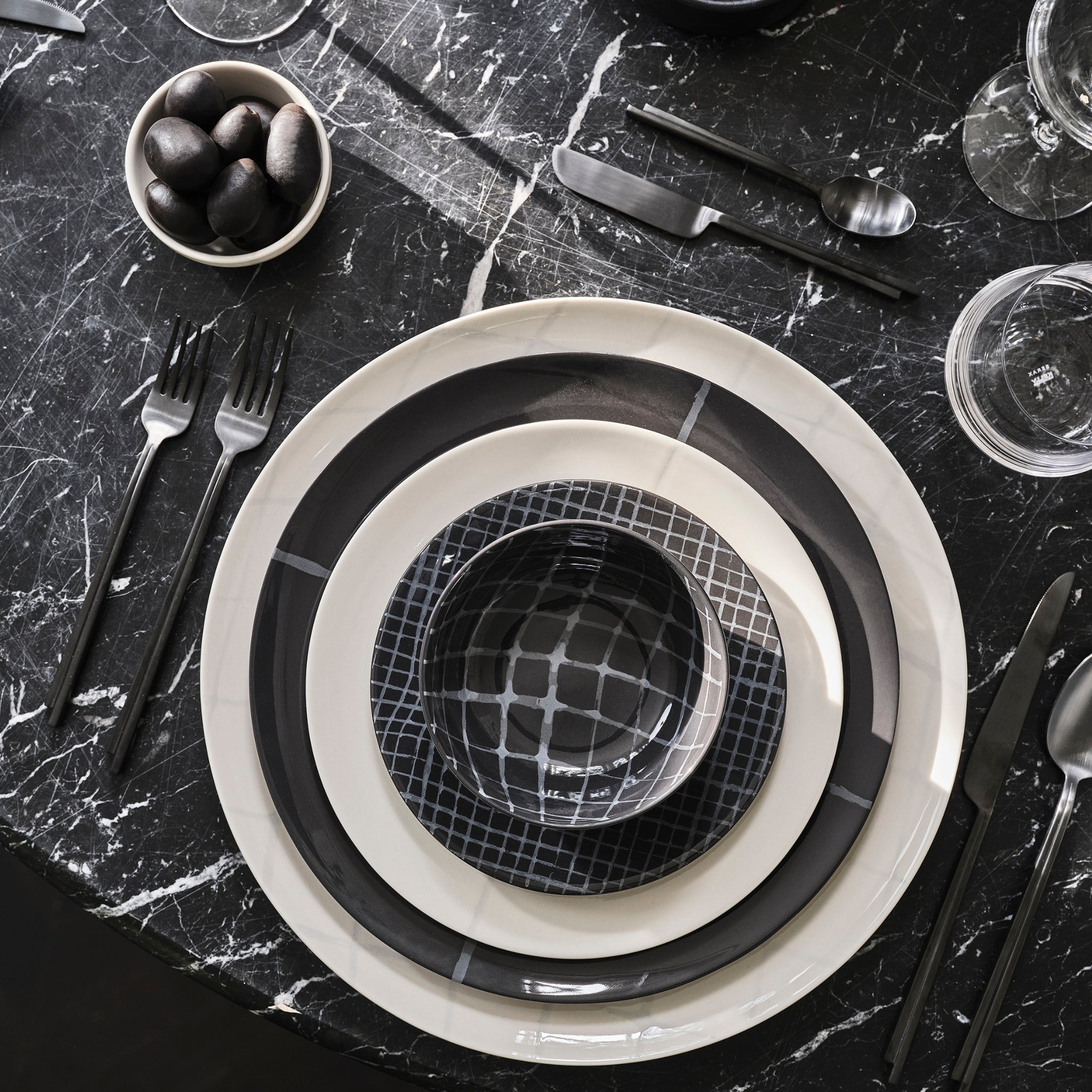 Een moderne, op Bauhaus geïnspireerde tafelsetting met de Serax Zuma Kom Pacific D12,5 kom van Serax, gestapelde zwarte en witte borden, matzwart bestek, zwarte olijven en heldere glazen op een donker marmeren oppervlak.