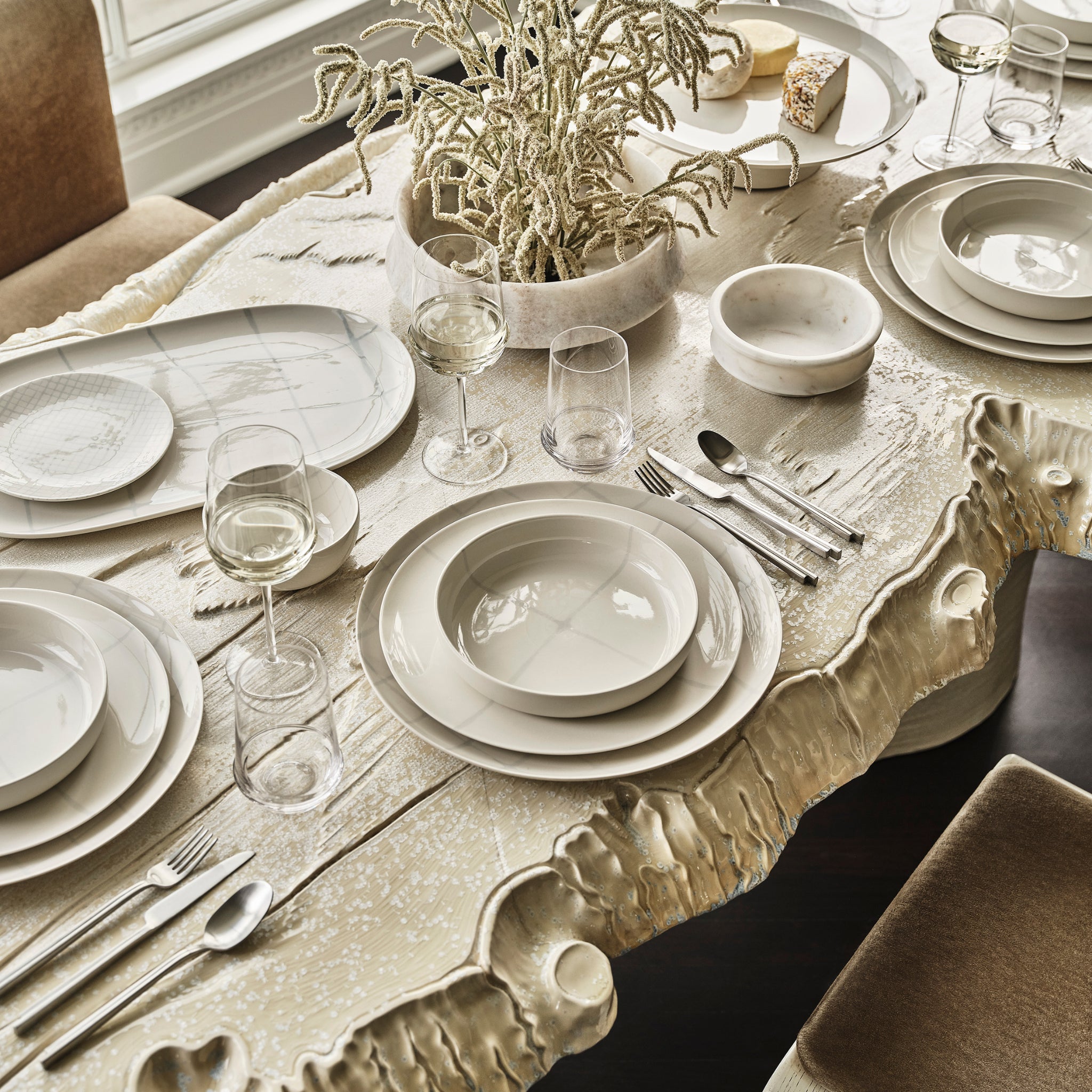 Een sculpturale crèmekleurige eettafel is elegant gedekt met Serax Zuma Kom Salt D19,5 kommen, vergezeld door neutraal getint servies en bestek. In het midden roepen decoratieve takken de door Kelly Wearstler geïnspireerde natuurlijke stijl op.