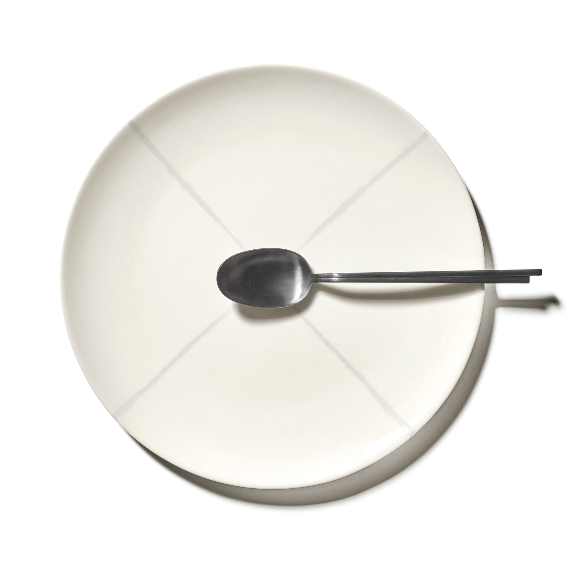 De Serax Zuma Laag Bord Salt D28, een effen wit bord met subtiele kruislijnen, wordt van bovenaf getoond met een zilveren lepel in het midden bovenaan, waarmee de minimale Bauhaus-geïnspireerde stijl van Serax wordt getoond tegen een witte achtergrond.