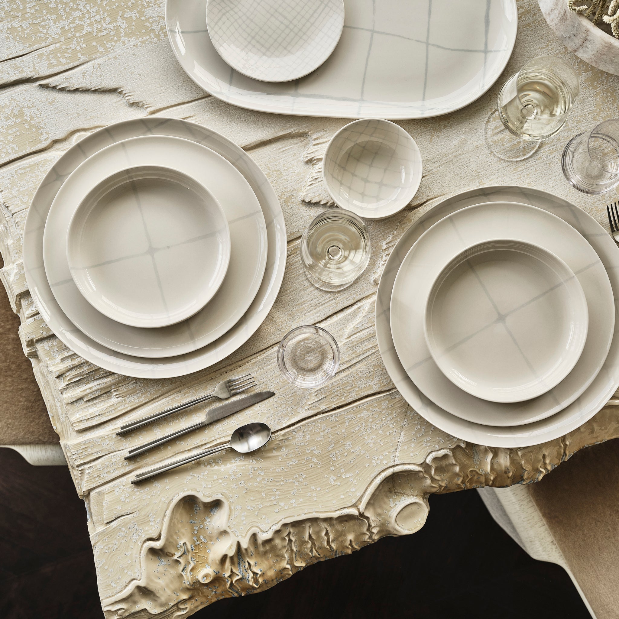 Een rustieke houten tafelset met de Serax Zuma Serveerschaal Salt D28,5 is voorzien van moderne witte borden met subtiele roosters, glaswerk en zilver bestek. De gestructureerde rand voegt een artistiek Bauhaus-geïnspireerd accent toe aan deze elegante Serax setting.