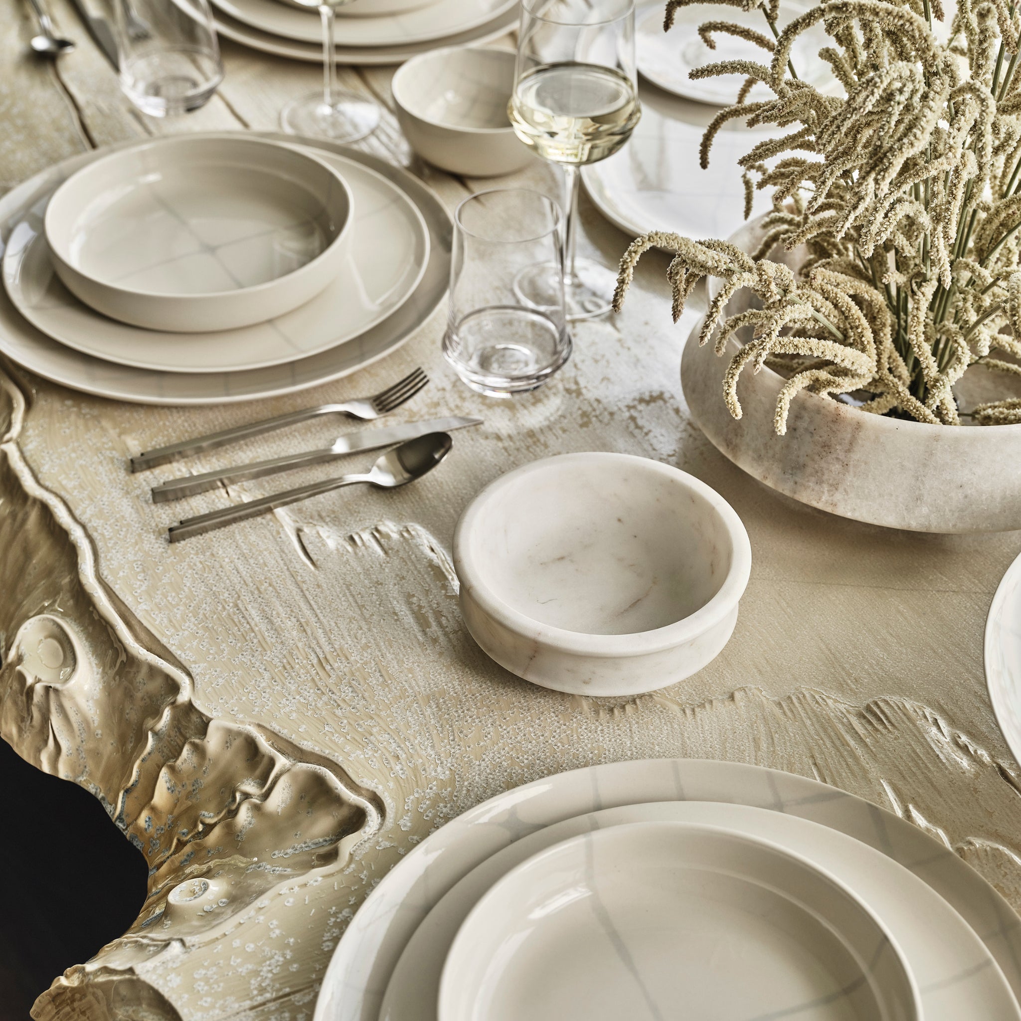 Een close-up van een elegante gedekte tafel met Serax Zuma Serveerschaal Salt D28,5-beige keramisch servies van Serax op een golvend gouden oppervlak, aangevuld met zilver bestek en een middenstuk van licht gedroogd gebladerte.