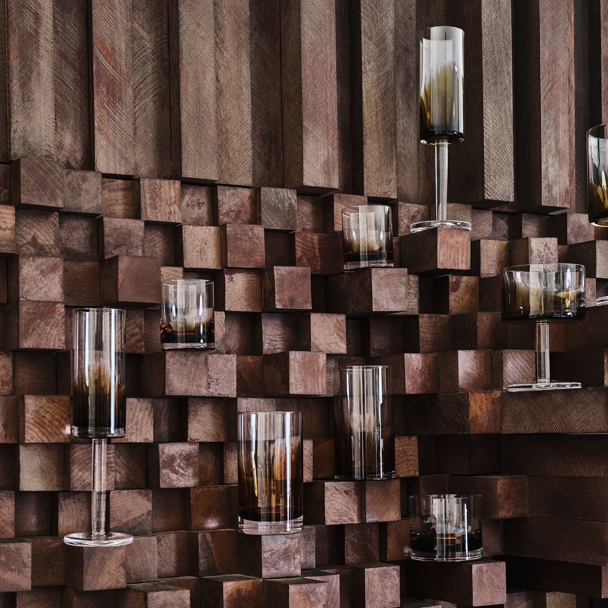 De Serax Zuma Whiskyglas Amber 20 cl van Serax, met een stijlvolle donker getinte basis, wordt tentoongesteld tegen een geometrische houten wand van vierkante houtblokken in verschillende bruintinten en dieptes.