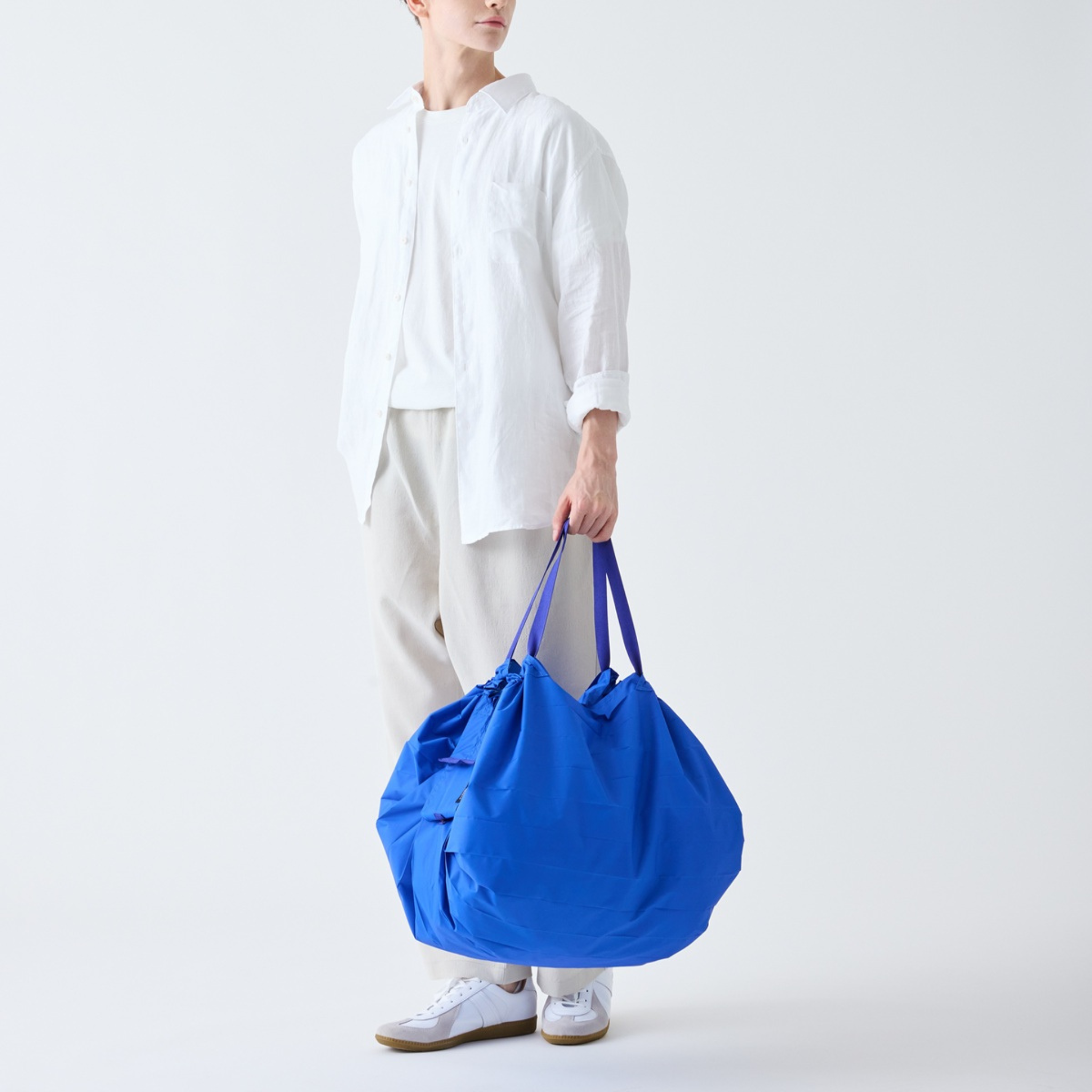 Een persoon in een los wit overhemd, een lichte broek en witte sneakers houdt een Shupatto Opvouwbare Boodschappentas Clear Sky Blauw L-draagtas met twee hengsels vast tegen een effen witte achtergrond.