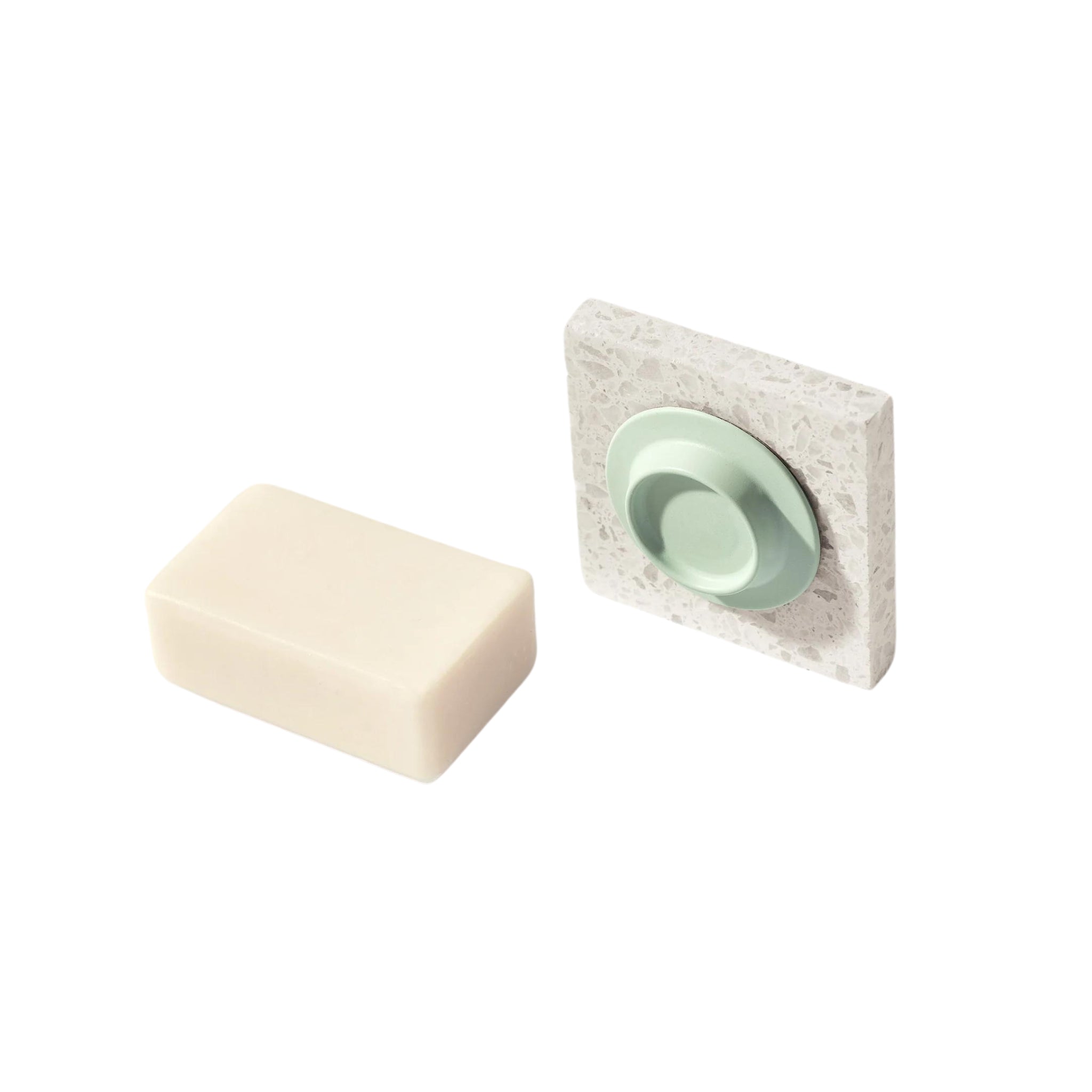 Een rechthoekig wit zeepstaafje ligt naast de Soapi Magnetische Zeephouder Munt, een vierkante, gespikkelde magnetische zeephouder met een rond, lichtgroen middenschaaltje van Soapi, beide op een strakke witte achtergrond.