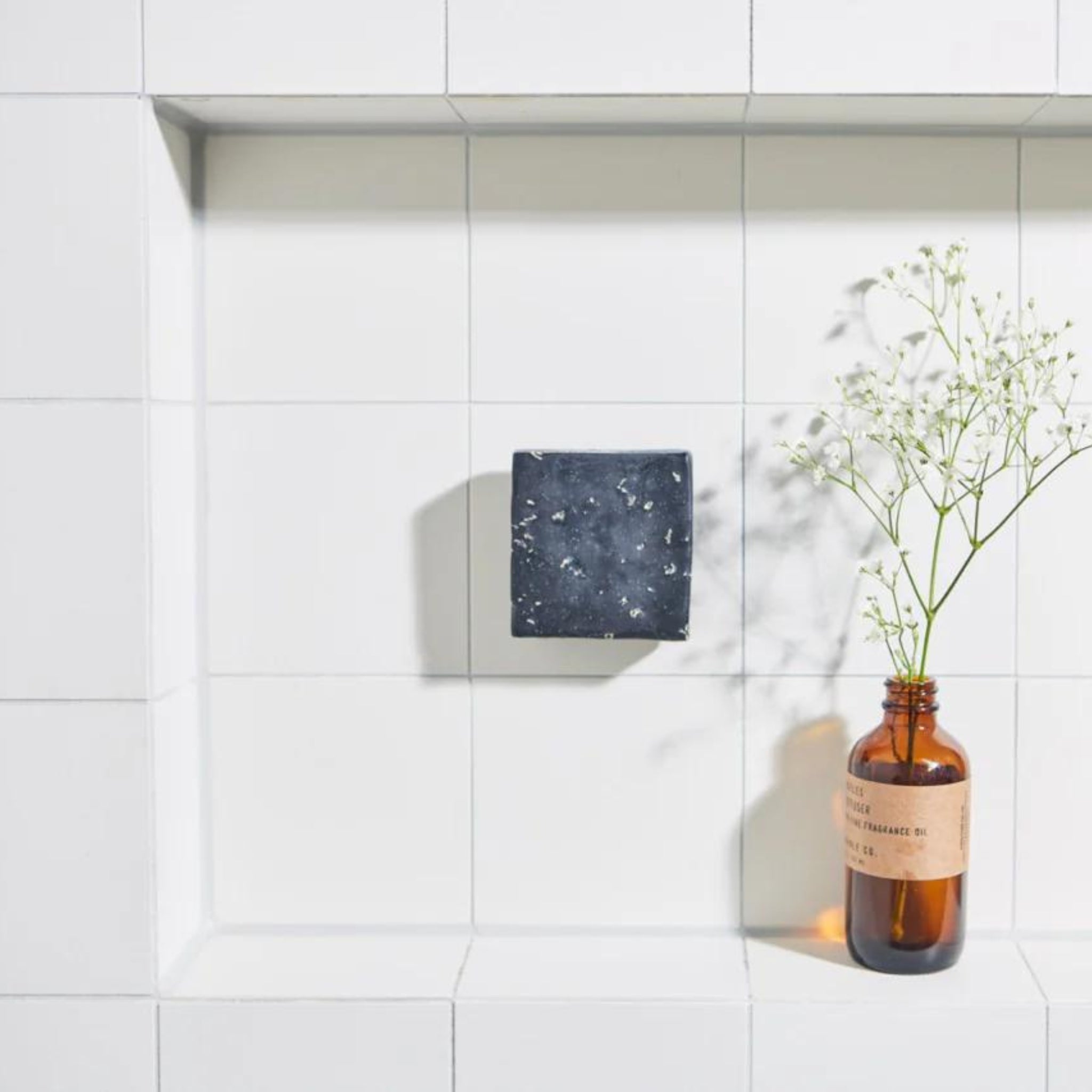 Een Soapi Magnetische Zeephouder Munt van Soapi in zwart met witte stippen rust op een wit betegeld plankje naast een bruine glazen fles gevuld met delicate witte bloemen.