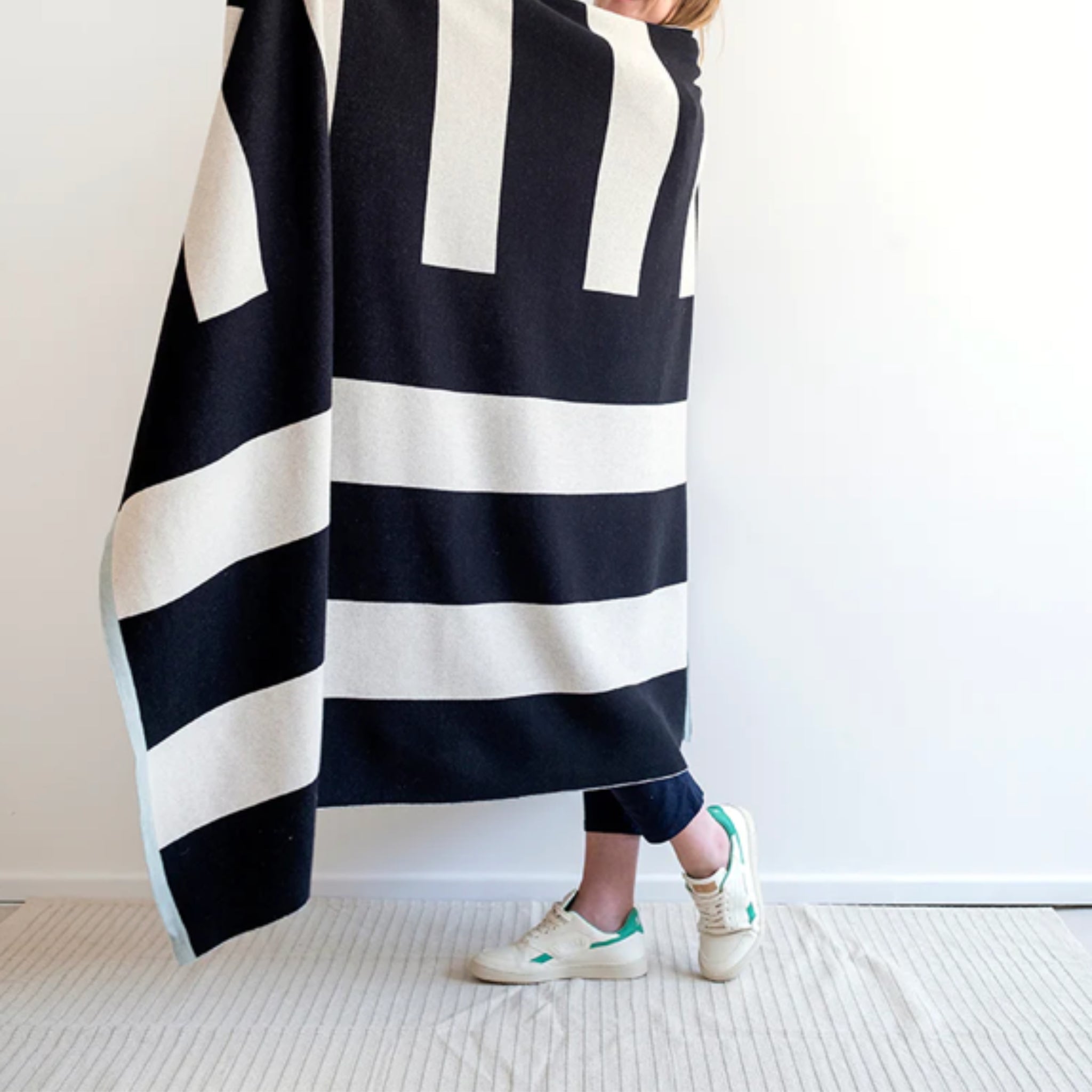 Een persoon gedeeltelijk bedekt door de Sophie Home Enkel Plaid Mono in zwart en wit staat op een licht vloerkleed in jeans en witte sneakers met groene accenten, tegen een effen witte muur die de stijlvolle interieuraccessoires van Sophie Home laat zien.