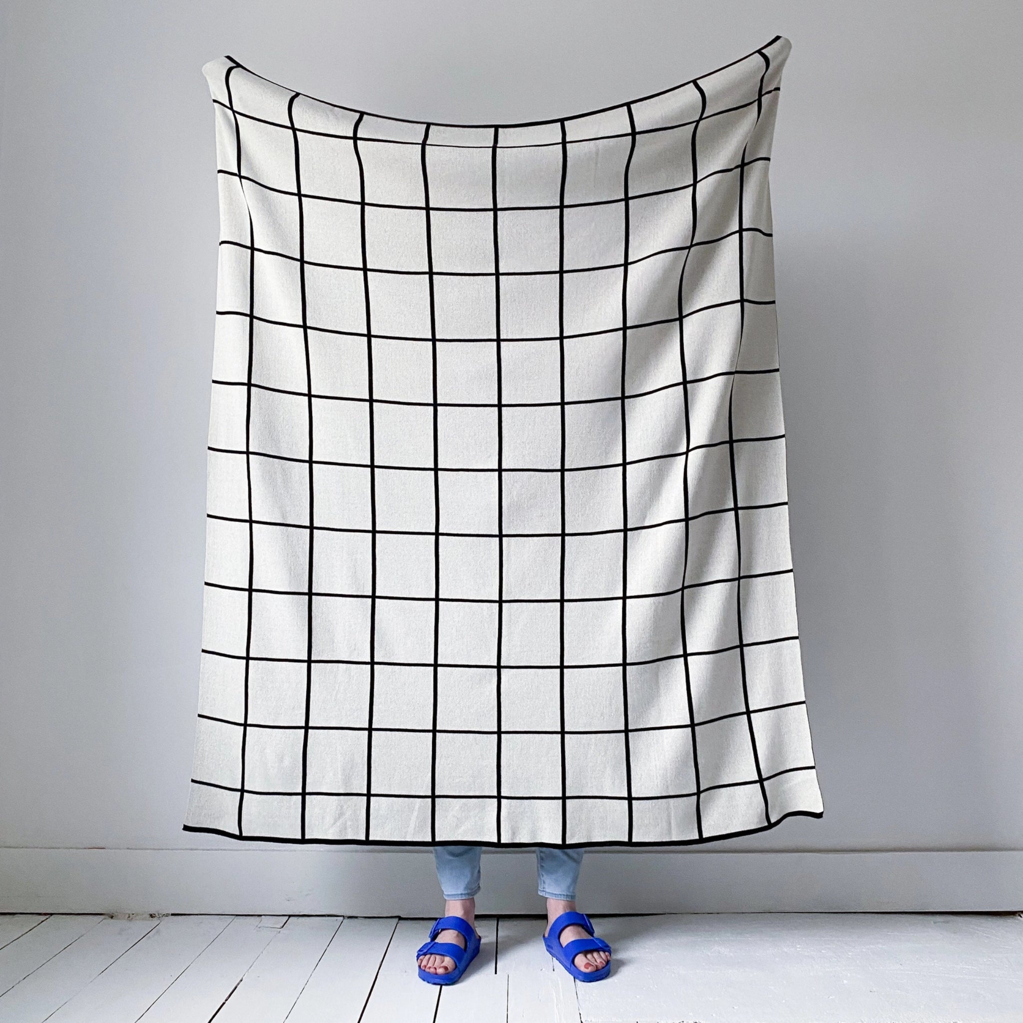 Een persoon staat achter de Sophie Home Grid Plaid Mono van Sophie Home, met alleen de onderbenen en voeten in blauwe sandalen en lichte spijkerbroek zichtbaar onder de deken. De achtergrond bestaat uit een effen witte muur en vloer.