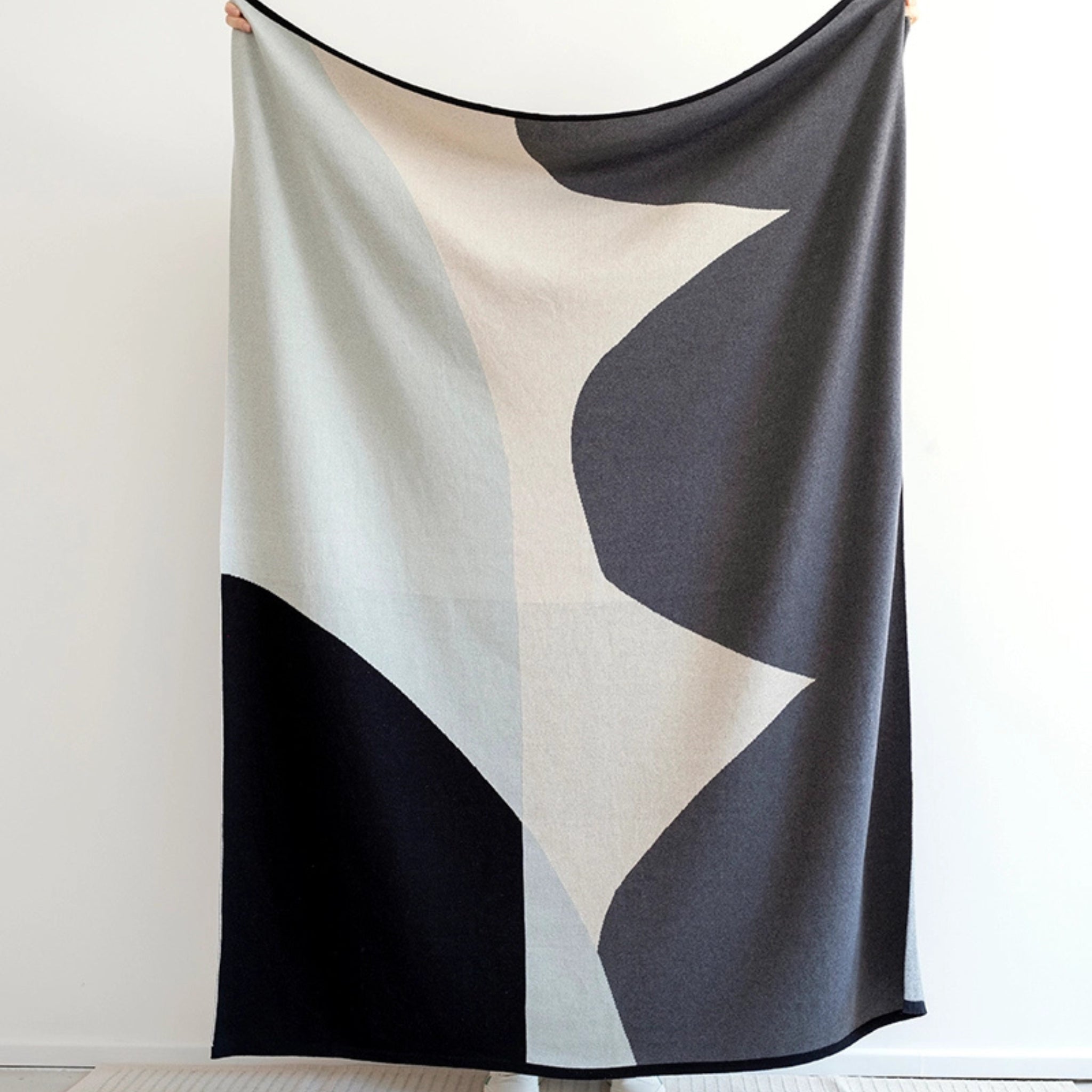 Persoon houdt een Sophie Home Stille Plaid Aqua van Sophie Home omhoog, met abstracte geometrische vormen in zwart, grijs en crème - ideaal als interieuraccessoires - tegen een effen witte achtergrond.