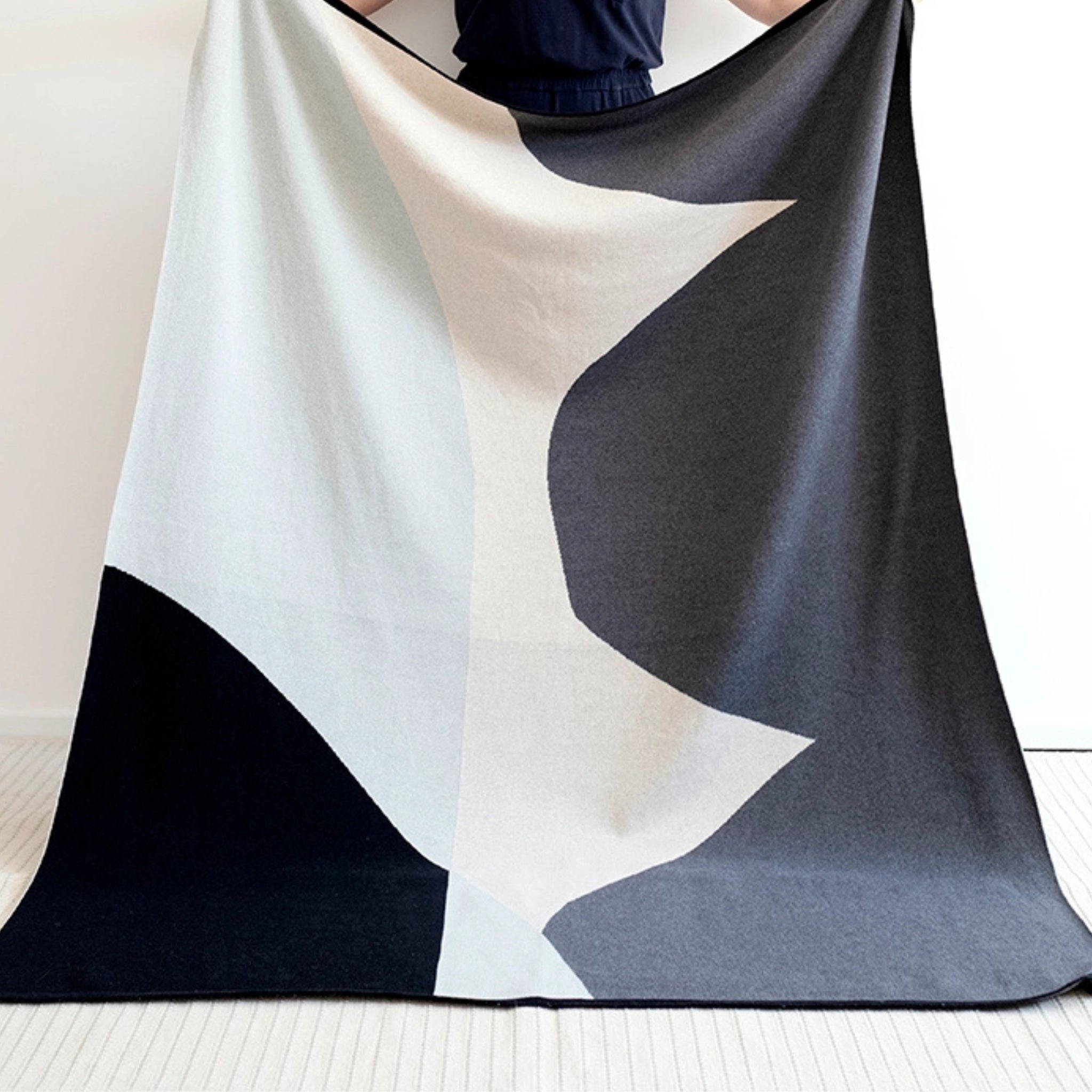 Een persoon houdt de Sophie Home Stille Plaid Aqua omhoog, een moderne deken van Sophie Home met abstracte geometrische vormen, waardoor dit stijlvolle accessoire opvalt tegen de effen witte muur en lichte vloer.