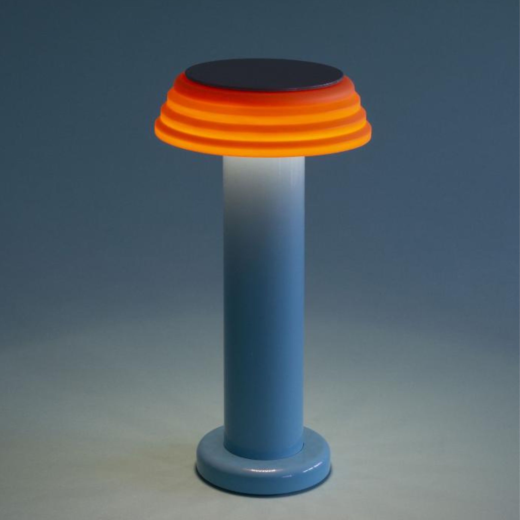 De Sowden PL1 Lamp LED Oplaadbaar Blauw van Sowden is een moderne tafellamp met een cilindrische blauwe voet en gelaagde oranjerode lampenkap, geïnspireerd op de Memphis Design Movement, die oplicht tegen een effen blauwe achtergrond.