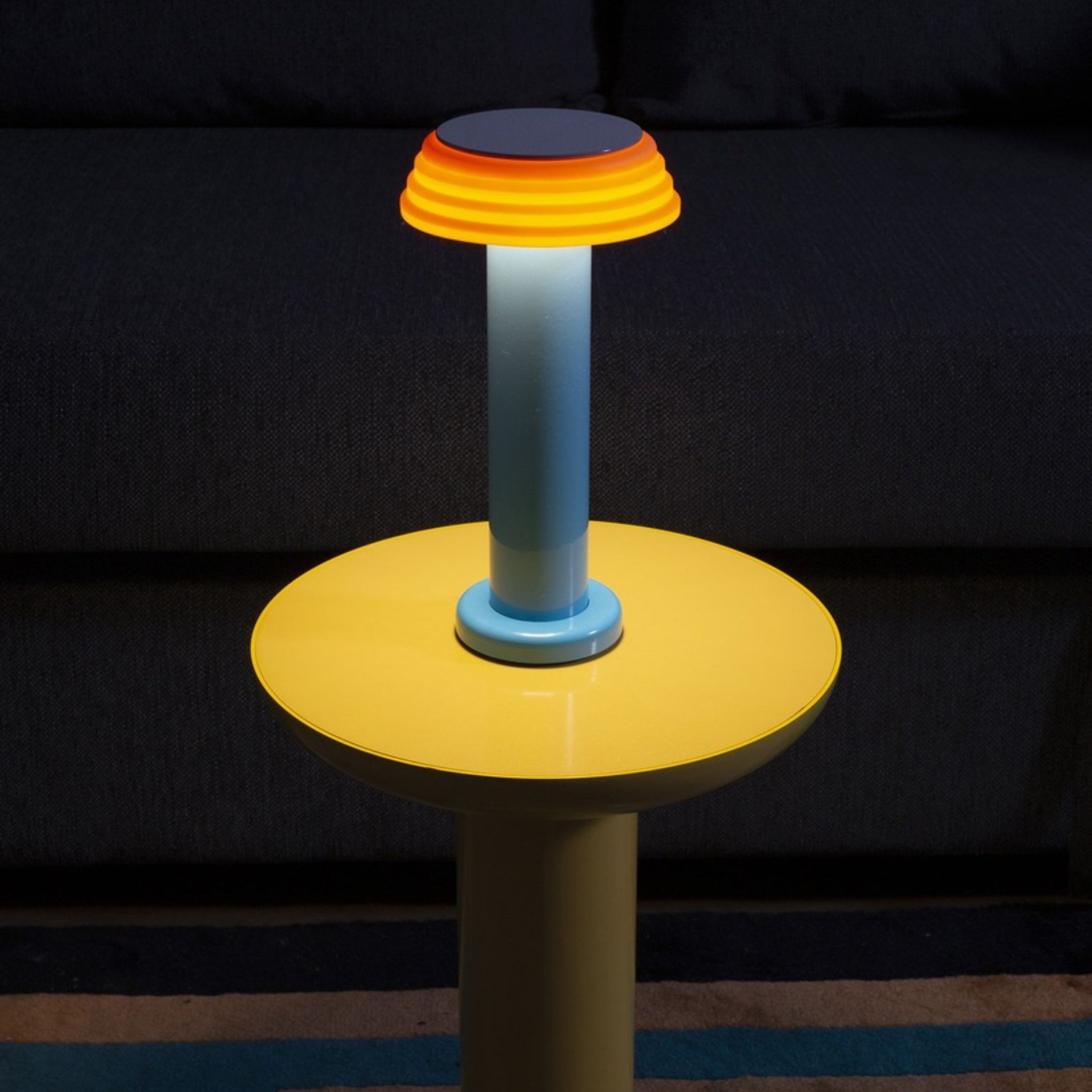 De Sowden PL1 Lamp LED Oplaadbaar Blauw van Sowden, met een blauwe voet en een gelaagde oranje-gele lampenkap geïnspireerd door de Memphis Design Movement, gloeit op een ronde gele tafel voor een donkere bank.