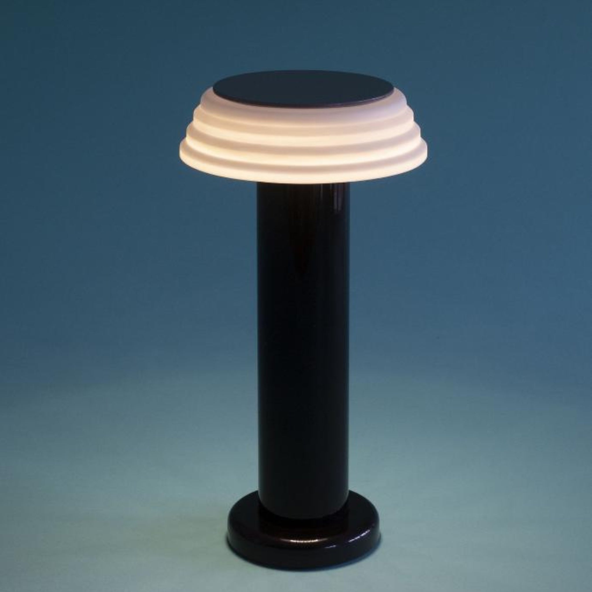 De Sowden PL1 Lamp LED Oplaadbaar Zwart van Sowden is een moderne, op Memphis geïnspireerde lamp met een cilindrische, zwarte voet en een witte, gelaagde, ronde kap die zacht licht uitstraalt tegen een blauwe achtergrond.