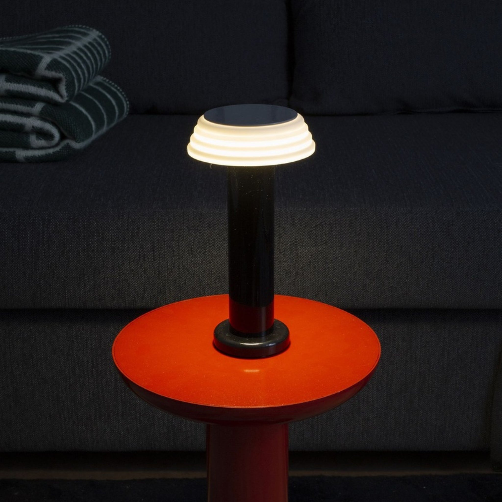 De Sowden PL1 Lamp LED Oplaadbaar Zwart van Sowden, geïnspireerd door de Memphis Design Movement, heeft een witte lampenkap en staat op een rond rood bijzettafeltje voor een donkere bank met een groen en wit deken op de achtergrond.