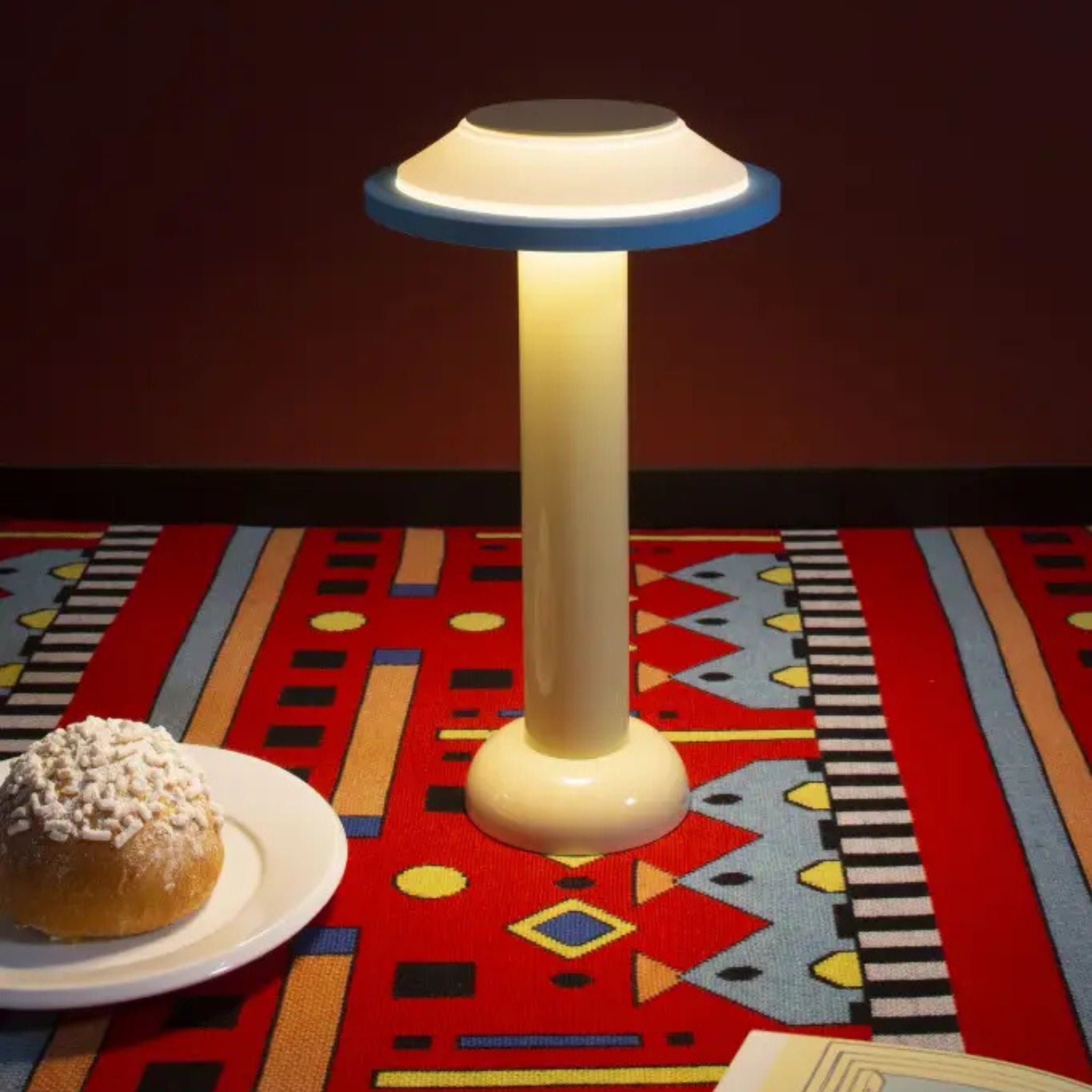 De Sowden PL2 Lamp LED Oplaadbaar Geel van Sowden werpt met zijn ronde kap een gloed op een kleurrijk geometrisch tafelkleed. Vlakbij rust een gepoederd gebakje op een wit bord tegen een donkerrode achtergrond.