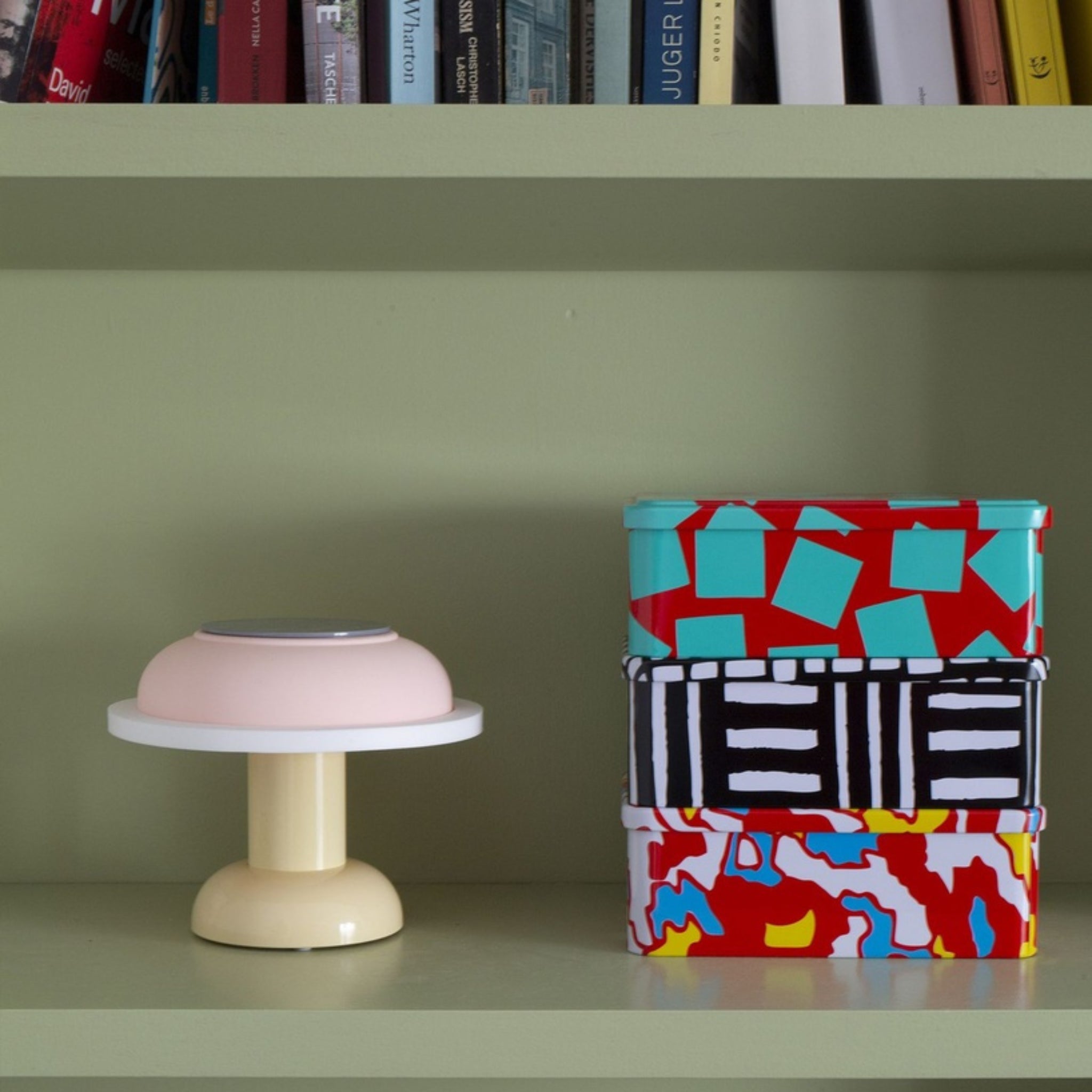 Op een lichtgroene plank staat een compacte, moderne Sowden PL4 Lamp LED Oplaadbaar Geel van Sowden naast twee blikken met een kleurrijk patroon. Boeken op de bovenste plank versterken de draadloze flair van draadloze verlichting.