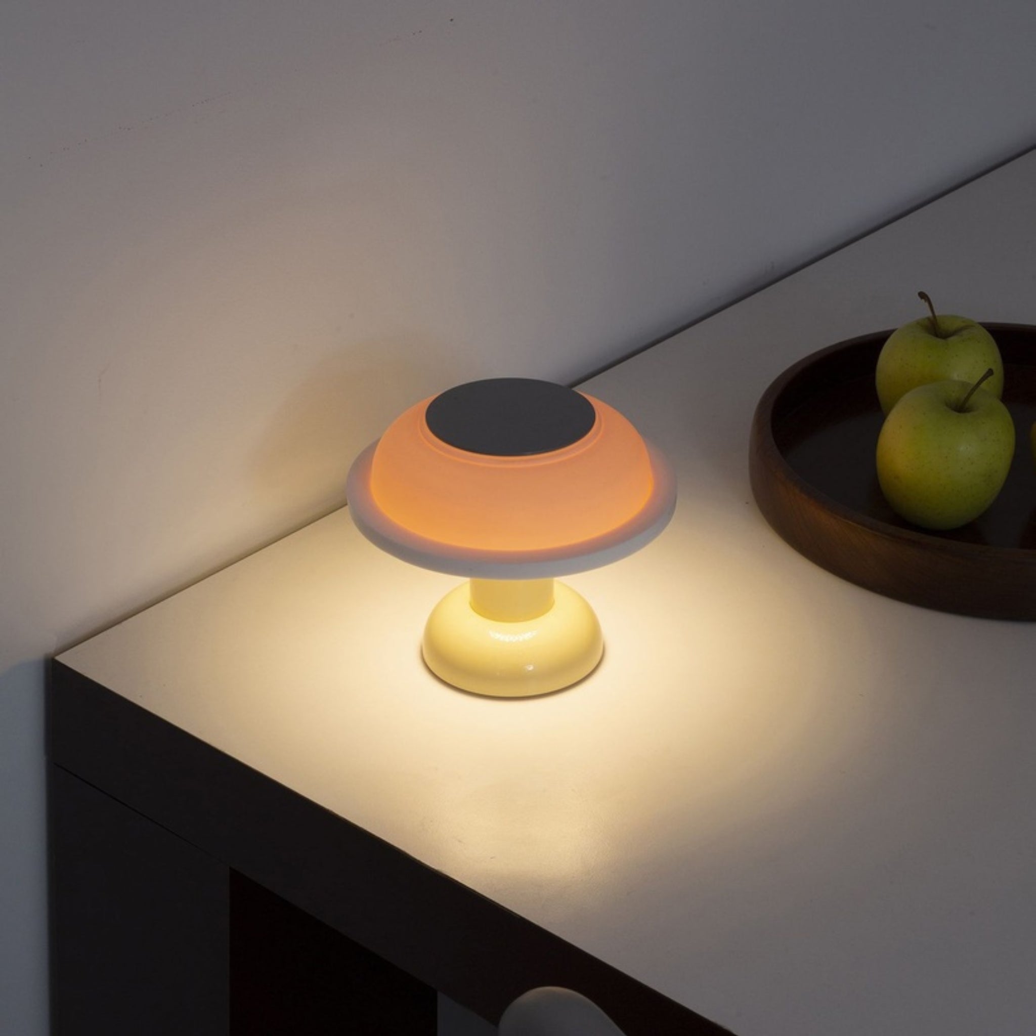 De Sowden PL4 Lamp LED Oplaadbaar Geel van Sowden, een moderne paddestoelvormige lamp, gloeit warm op een wit tafelblad naast een bruin dienblad met twee groene appels-een minimalistisch tafereel dat draadloze verlichting benadrukt.