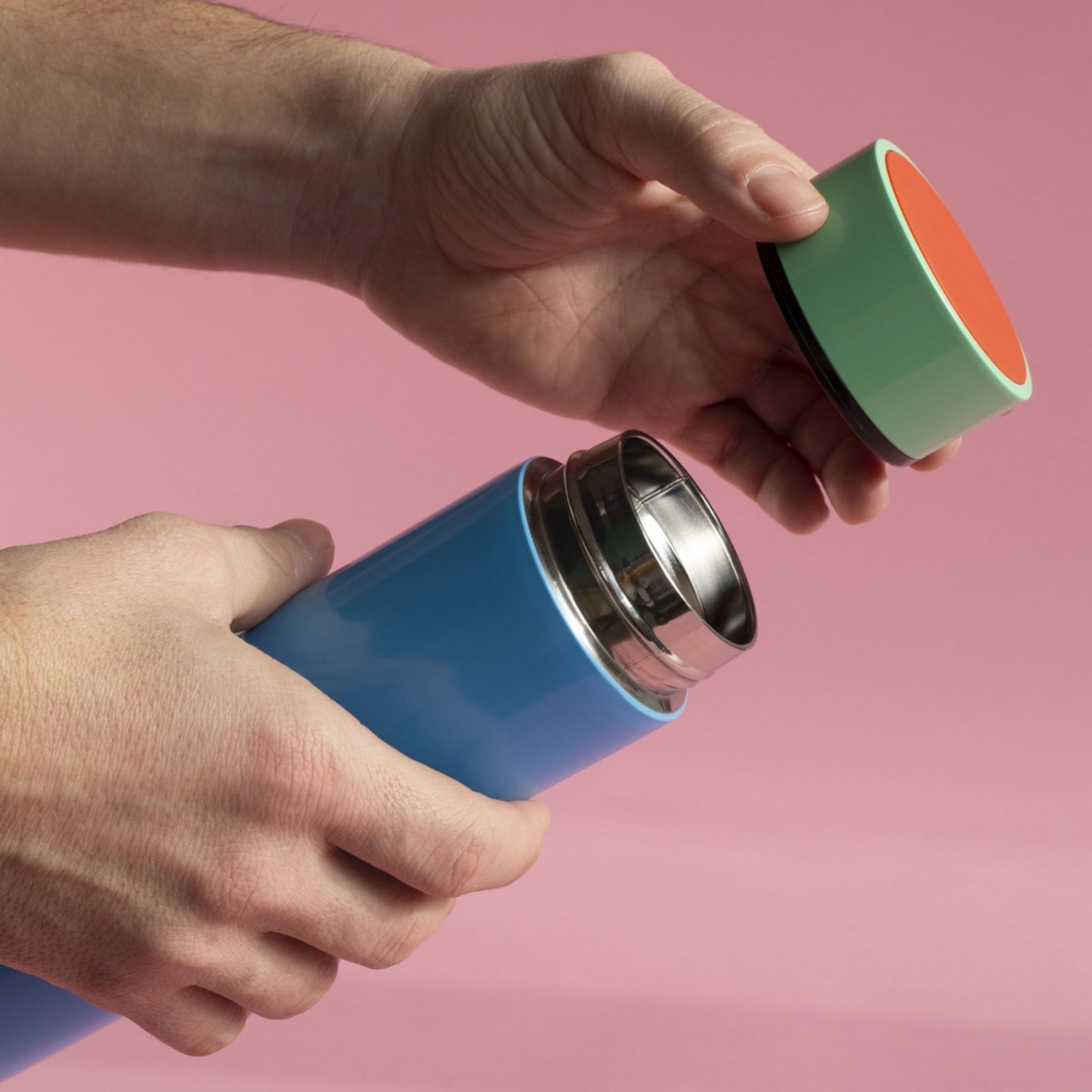 De handen van een persoon openen een Sowden Thermosfles Blauw 400 ml met een groen en oranje deksel tegen een roze achtergrond.