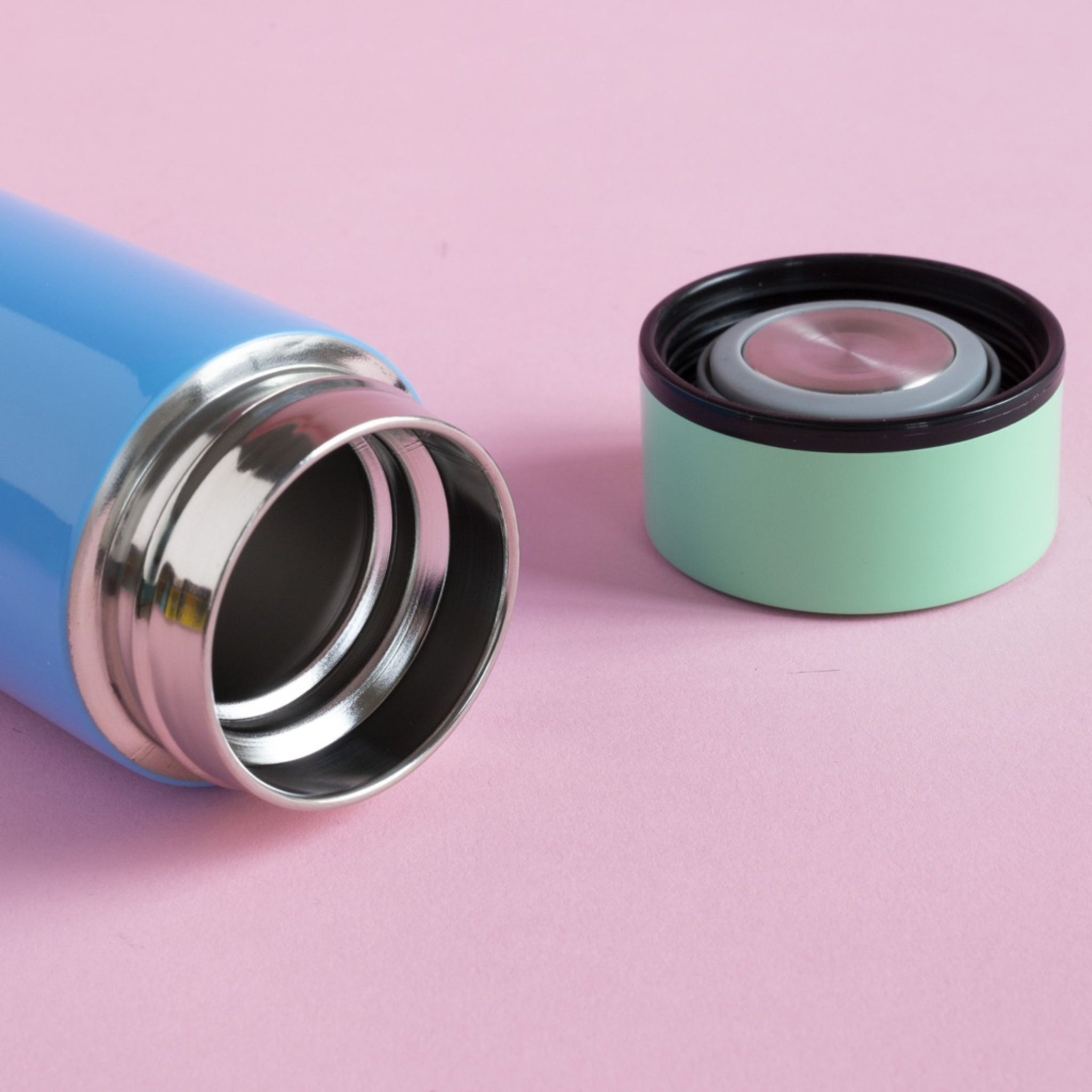 De Sowden Thermosfles Blauw 400 ml van Sowden rust op zijn kant naast zijn groen met zwarte deksel, ontworpen door George Sowden, op een roze achtergrond.